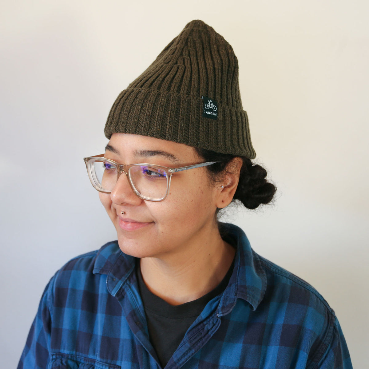A person with light brown skin and glasses wearing a dark green Tandem Coffee Roasters merino wool beanie and a blue plaid shirt, smiling slightly and looking to the side against a light background.