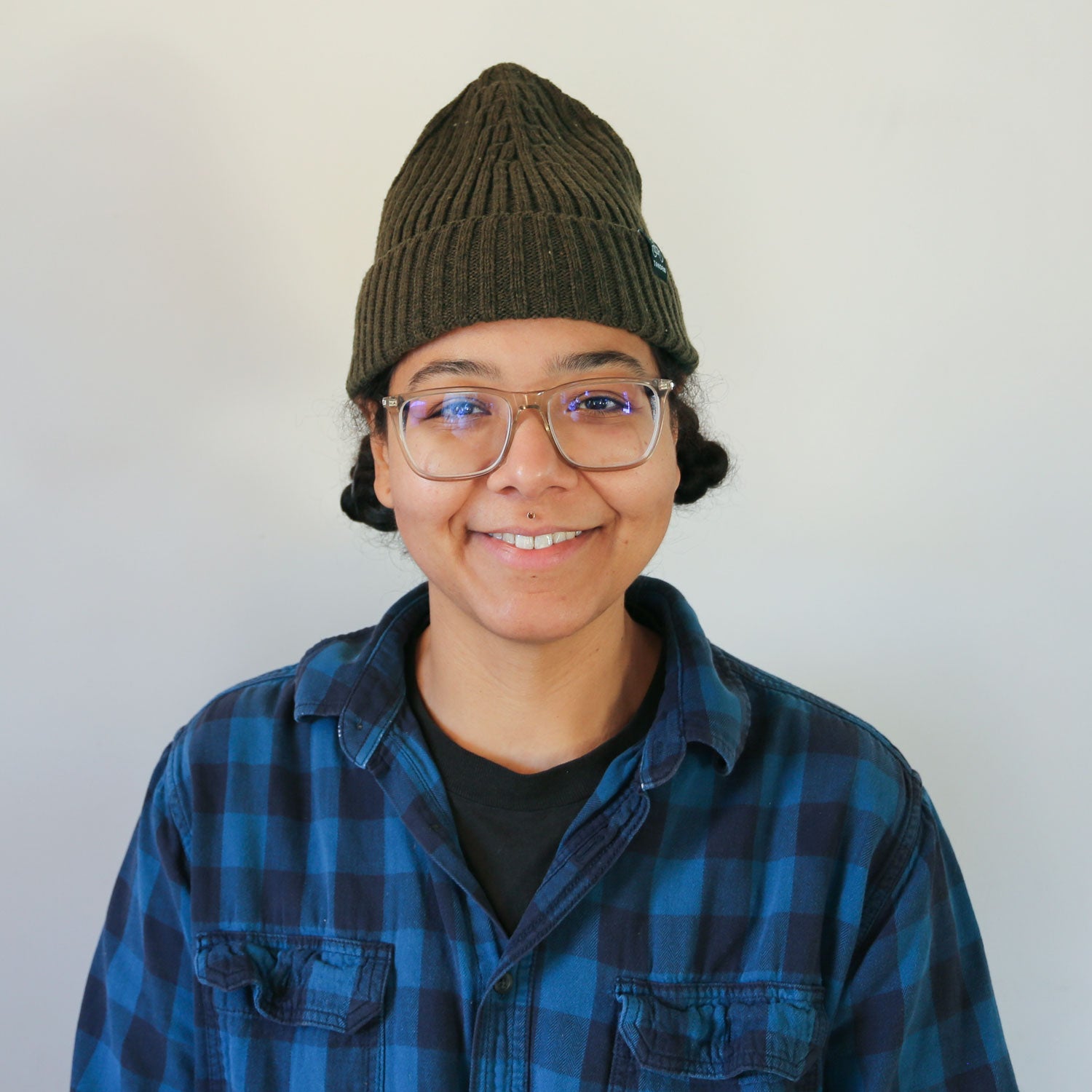 A young person wearing a green Tandem Coffee Roasters merino wool beanie, glasses, and a blue plaid shirt smiles warmly at the camera, against a light neutral background.
