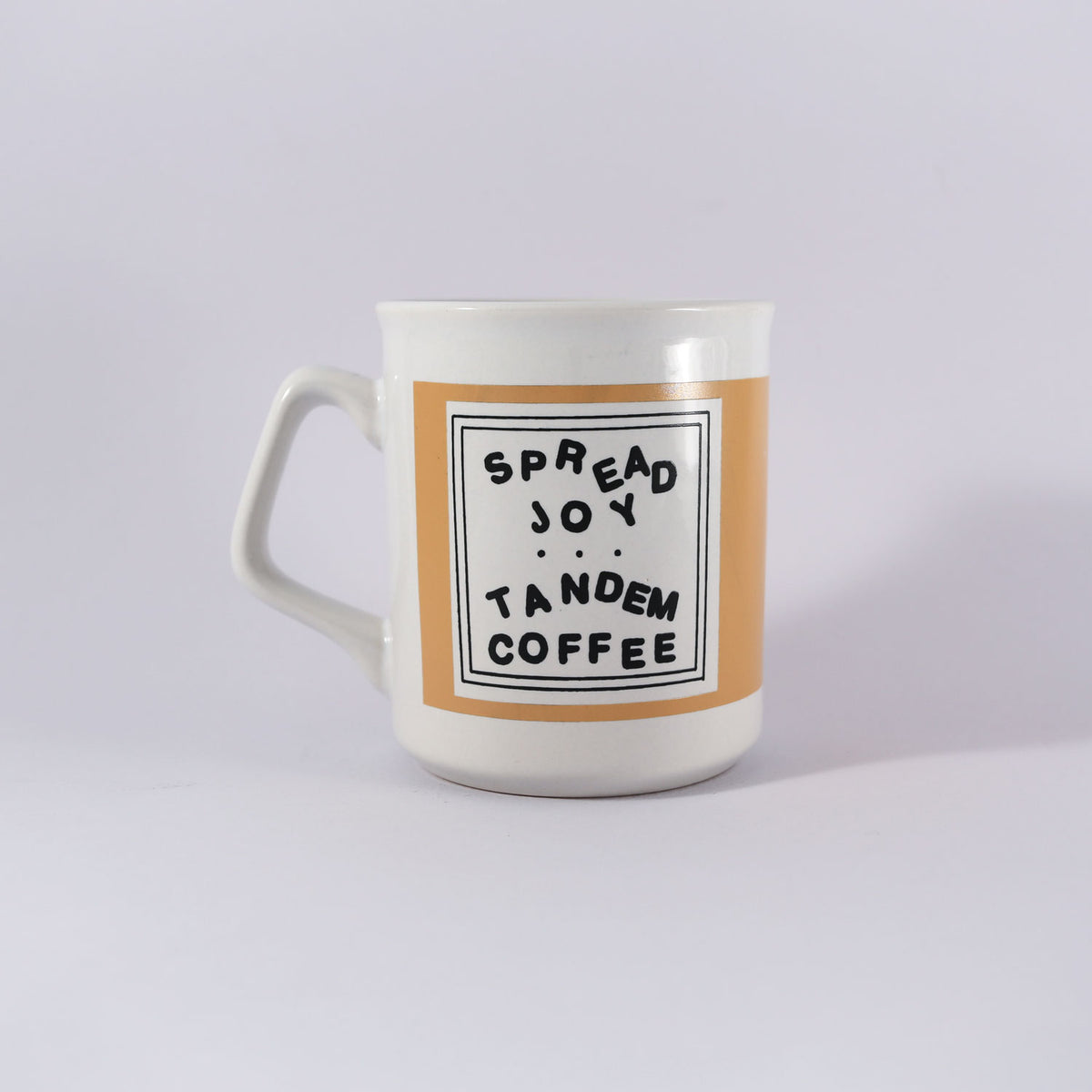 A white ceramic mug with a beige center band featuring the text "Kathy Heideman Spread Joy Mug" in a black square frame on a plain background, recommended for handwash only. Made by Tandem Coffee Roasters.