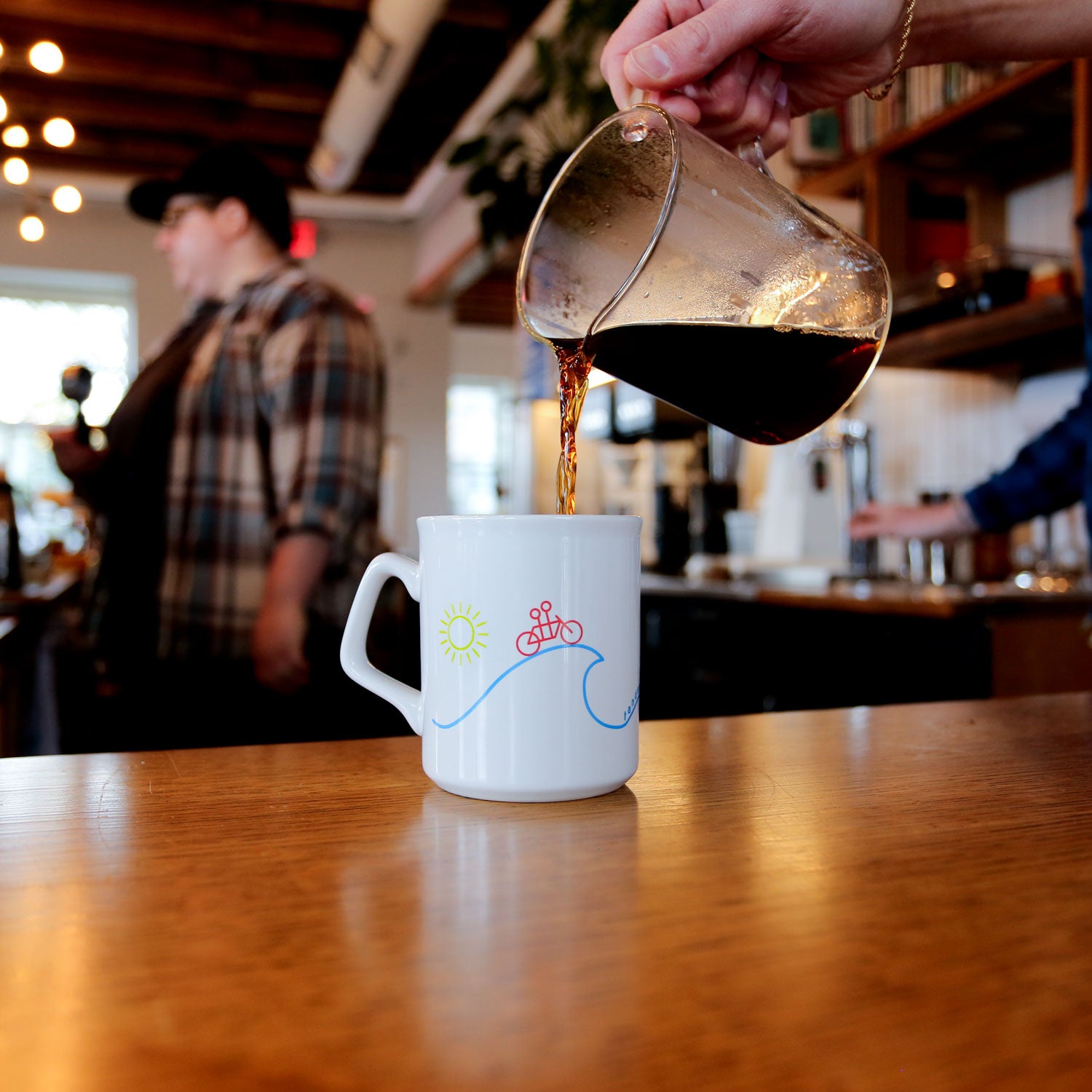 Coffee being poured into a Tandem Coffee Roasters Sunshine Mug with a bicycle and sun design on it in a cozy café setting.