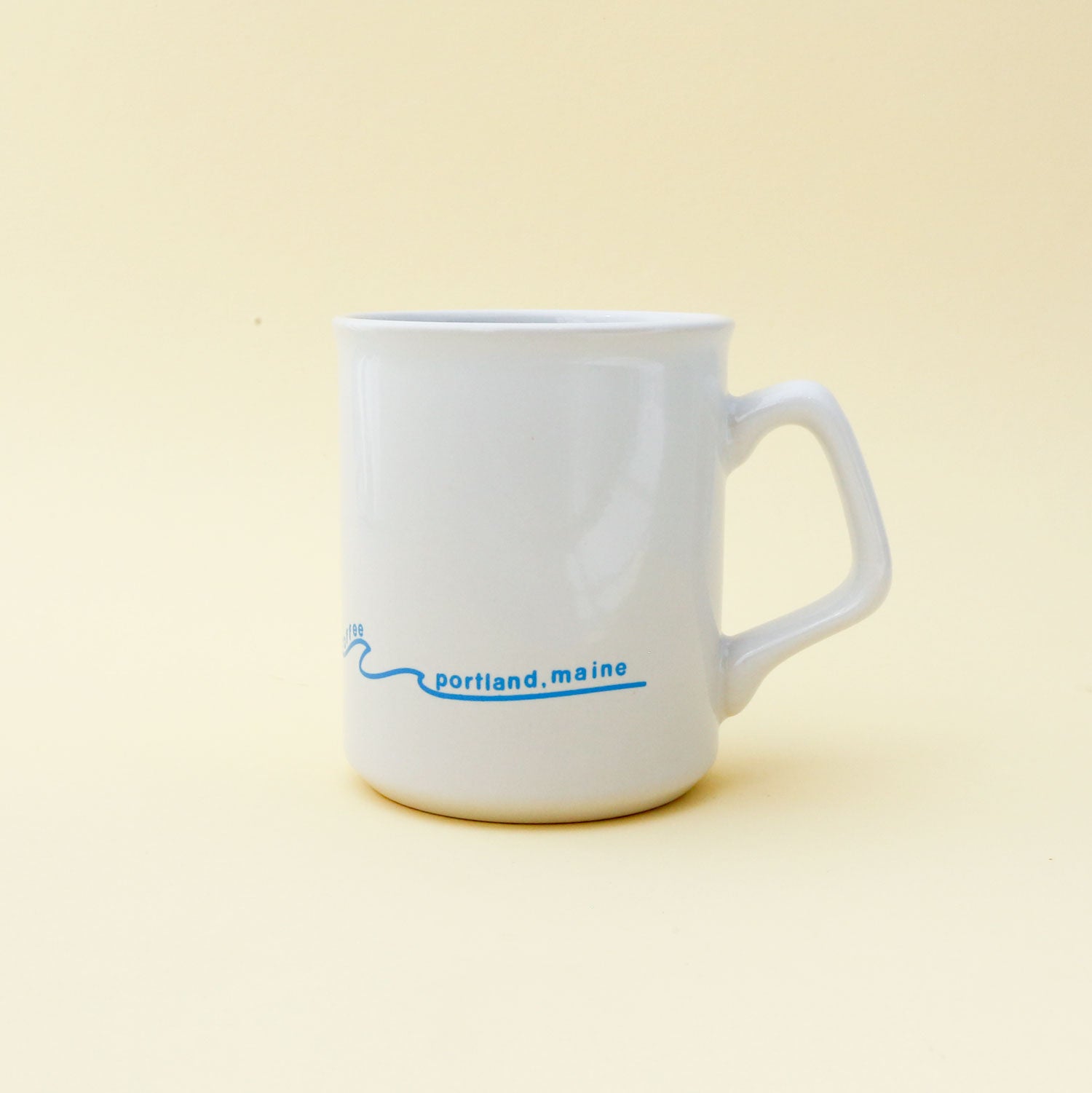 A white Sunshine Mug with a blue logo and text that reads "Portland, Maine" on a pale yellow background. The logo includes a small, stylized blue wave by Tandem Coffee Roasters.