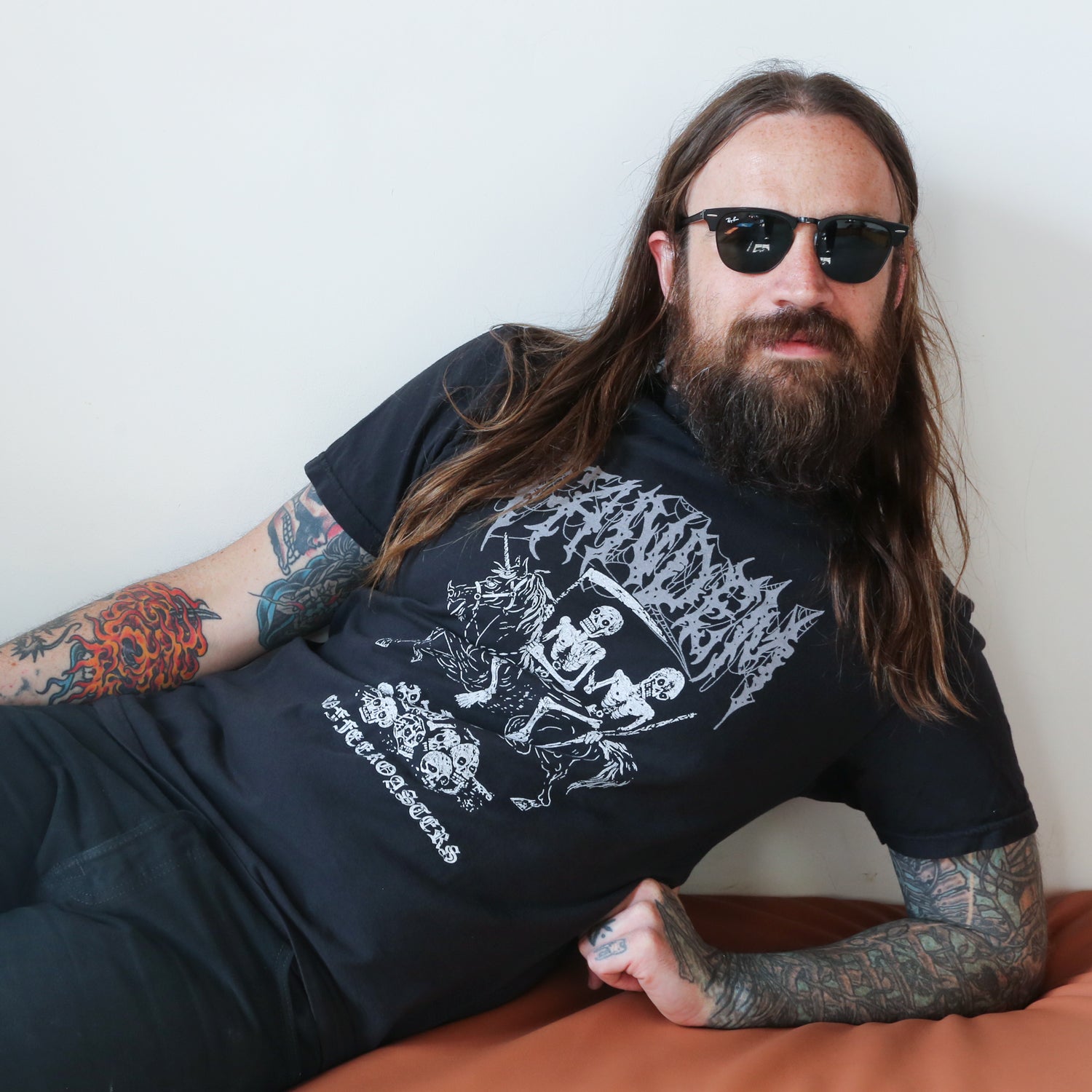 A man with long hair and a beard, wearing sunglasses and a Tandem Metal Tee from Tandem Coffee Roasters with graphic print, reclines on a beige sofa. His arms, featuring colorful tattoos including a prominent gold star tattoo.