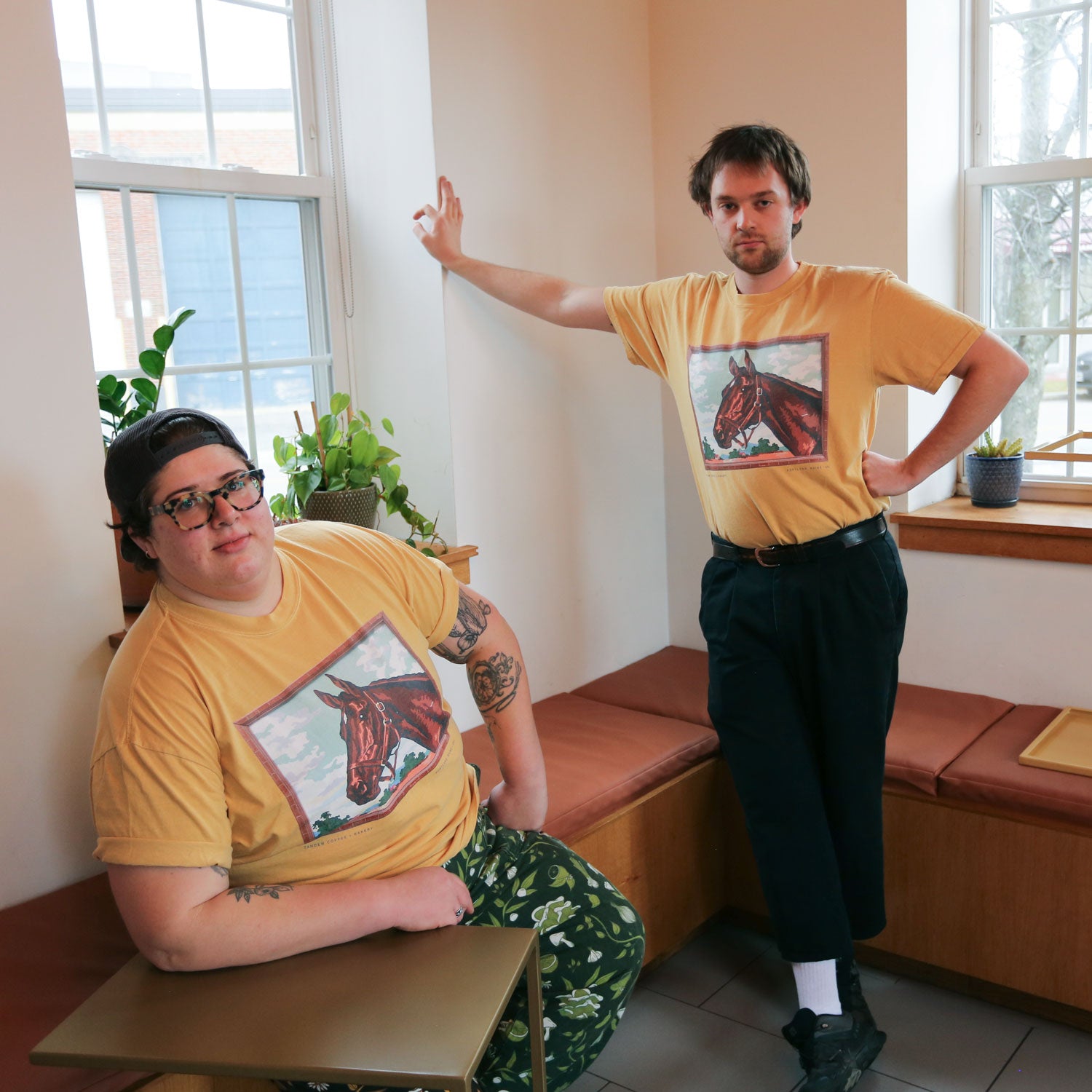 Two people wearing matching Tandem Coffee Roasters Paint by Number Bakery Horse tees pose in a brightly lit room with plants and benches; one is sitting on a desk, the other standing with a hand on the wall.