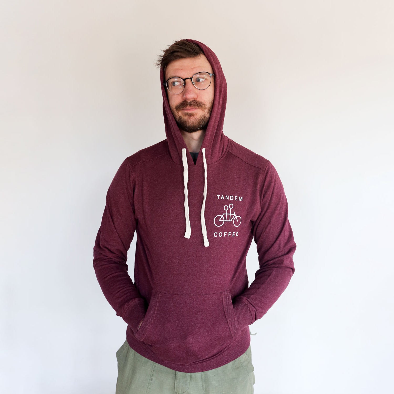 A man in glasses wearing a Maroon Recycled Pullover Hoodie from Tandem Coffee Roasters stands against a light grey background, looking at the camera with a slight smile.