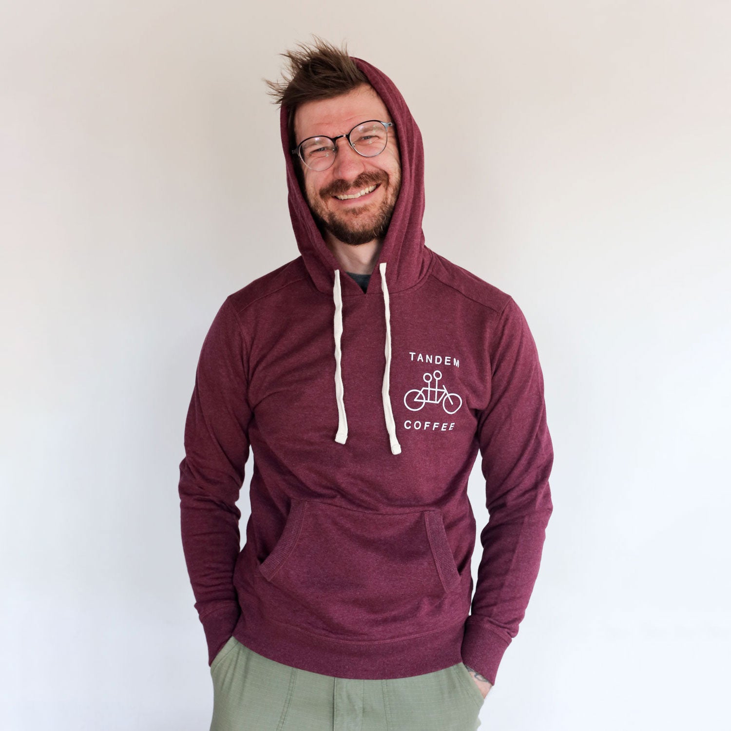 A man in glasses wearing a Maroon Recycled Pullover Hoodie from Tandem Coffee Roasters stands against a light grey background, looking at the camera with a slight smile.