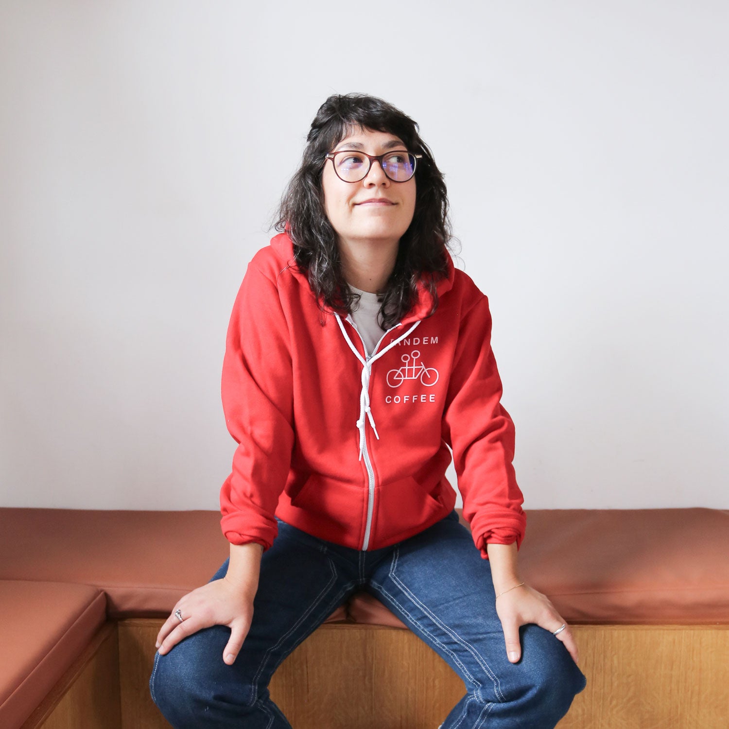 A young woman with glasses and curly hair, wearing a red Tandem Coffee Roasters zip-up hoodie, sits on a brown bench, looking upwards with a subtle smile.