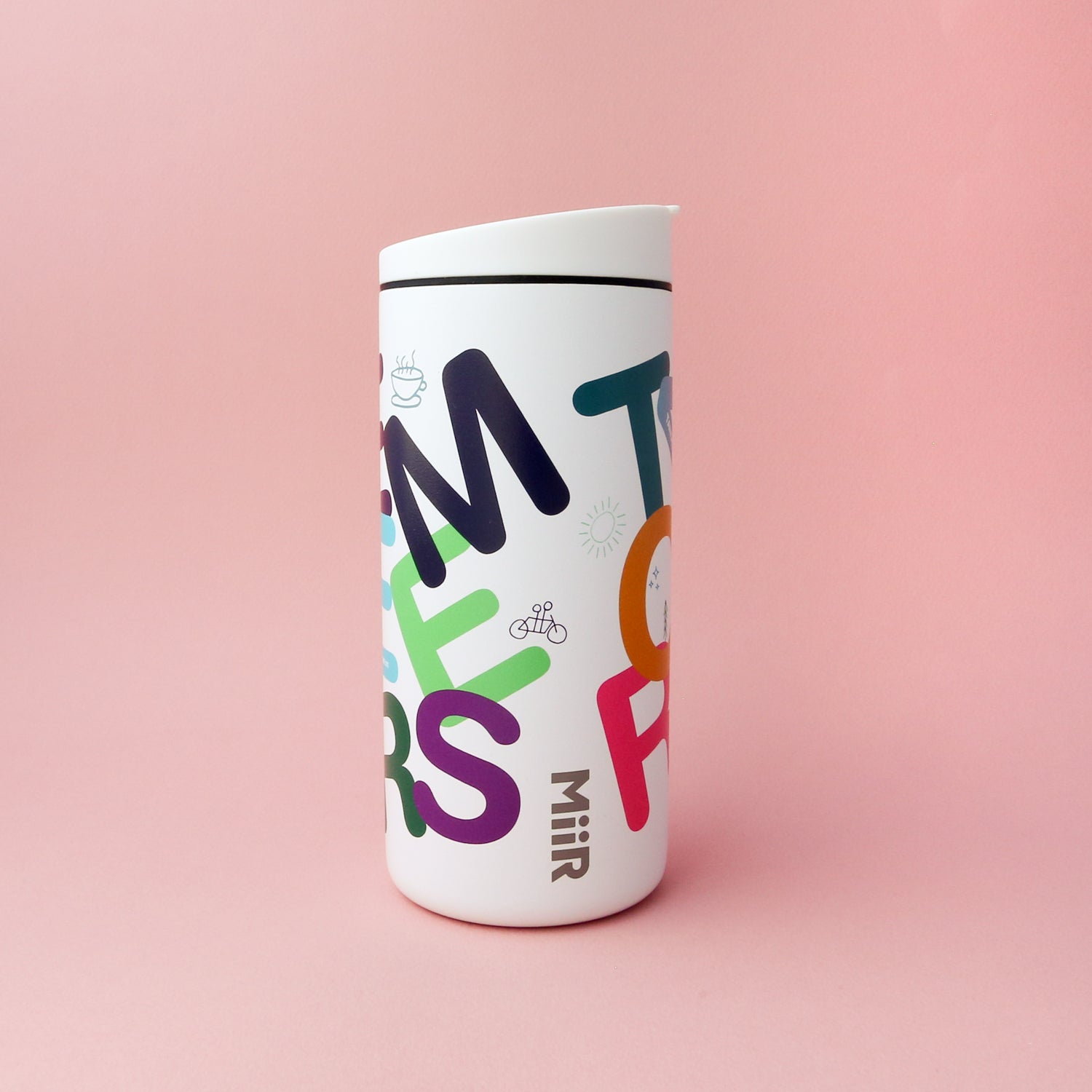 A white Letter Mug with playful, multicolored text and small illustrations scattered across the surface. Featuring a leak-proof lid, double wall vacuum insulation, and the words "Tandem Coffee Roasters" written vertically near the bottom, it stands out against a solid pastel pink background.