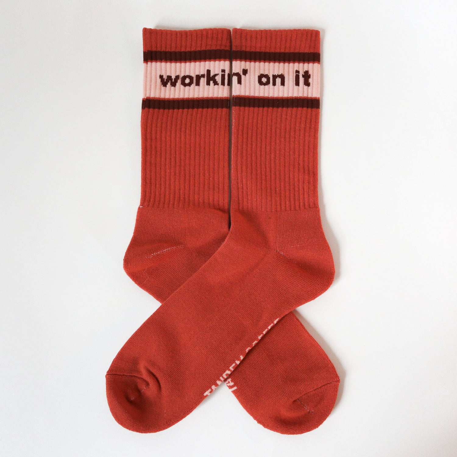 Introducing The Tandem Socks by Tandem Coffee Roasters: a lively pair of red cotton socks with pink bands at the top, showcasing the phrase "workin' on it" in dark red. Neatly crossed and set against a pristine white background, these socks offer a refreshing touch akin to enjoying morning coffee at Tandem Coffee.