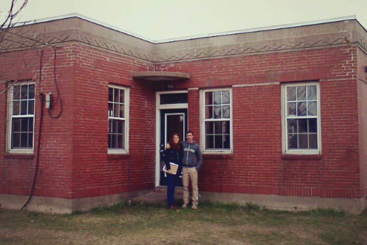 Two people, one holding a folder, standing in front of a red brick building with a classical design, featuring symmetrical windows and a central door.