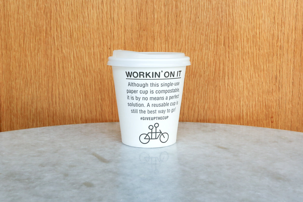 A white disposable coffee cup with the text "workin' on it" and an eco-friendly message on a marble table against a wooden panel background.