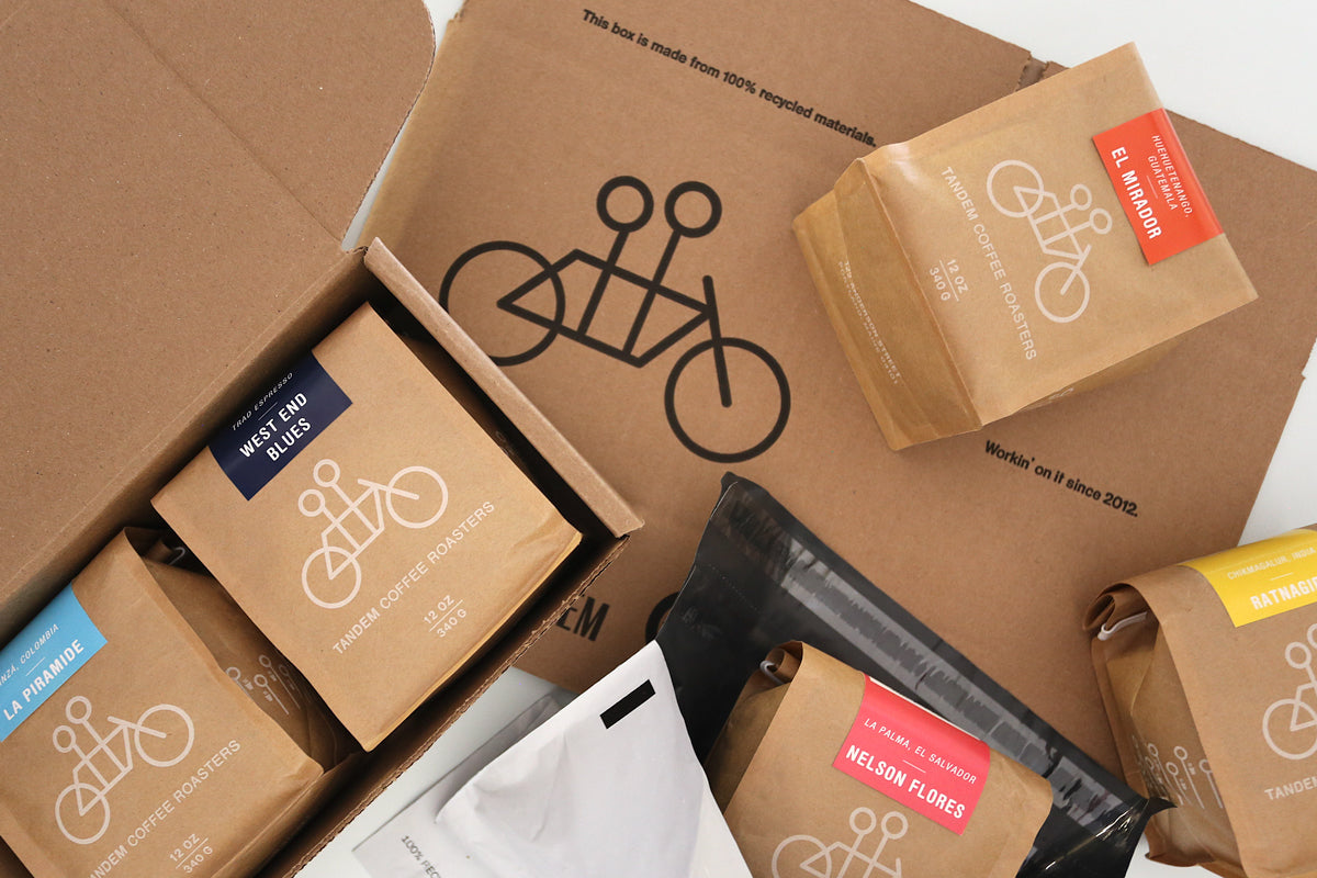 A top-down view of an open cardboard box filled with various neatly arranged packages and food products branded with bicycle logos, suggesting a delivery focused on cycling or environmental sustainability.