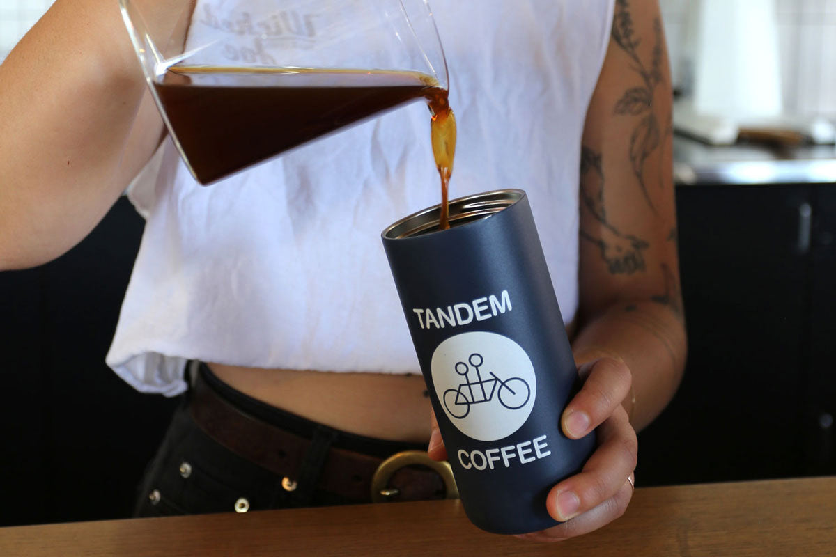 A person pours coffee into a dark blue travel mug branded with "tandem coffee" featuring a bicycle logo, held in a tattooed hand. only the person's torso and arms are visible.
