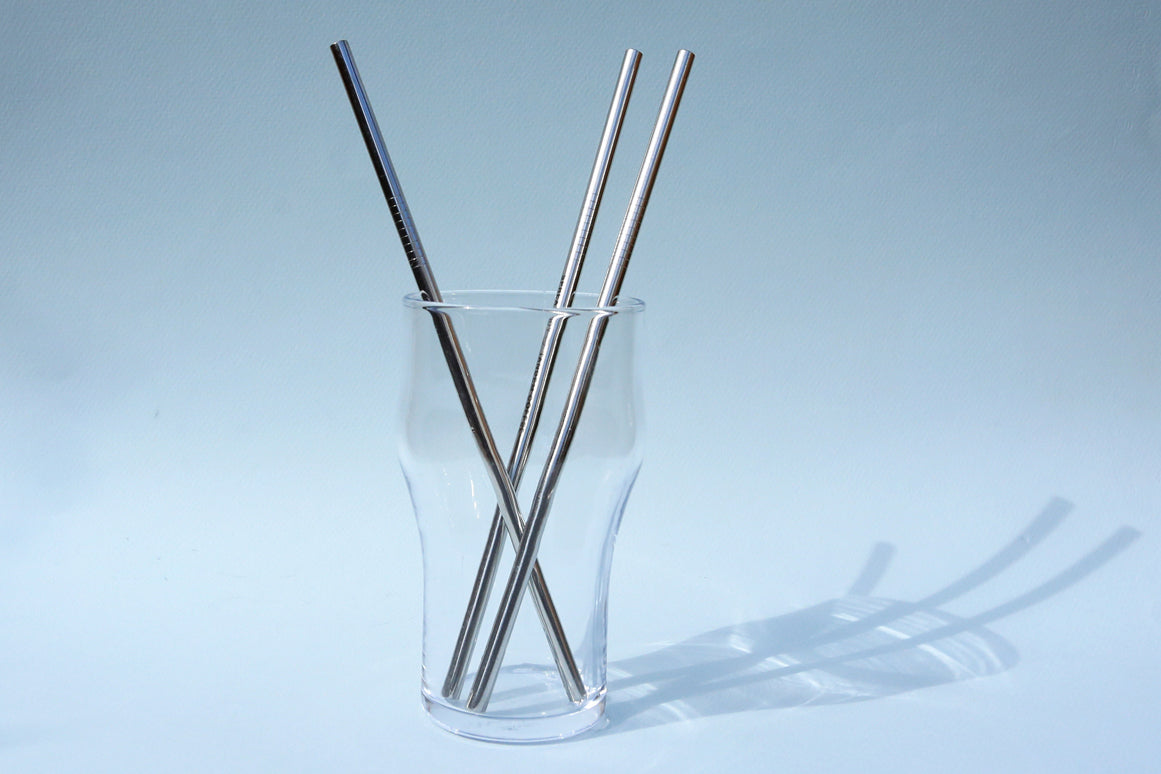 Three metal straws—two silver, one black—in a clear glass against a soft blue background, casting elongated shadows.