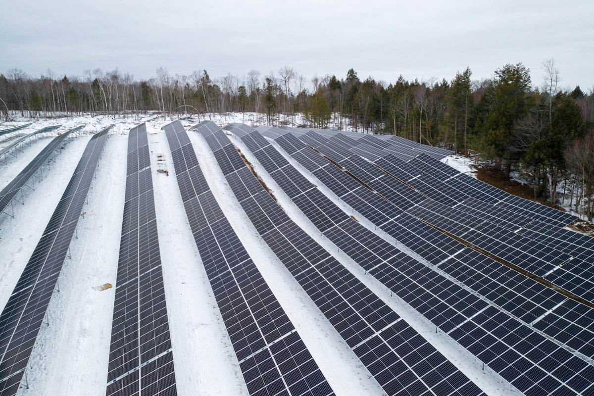 Aerial view of a solar farm with rows of solar panels partially covered in snow, amidst a forested area displaying a wintry scene.