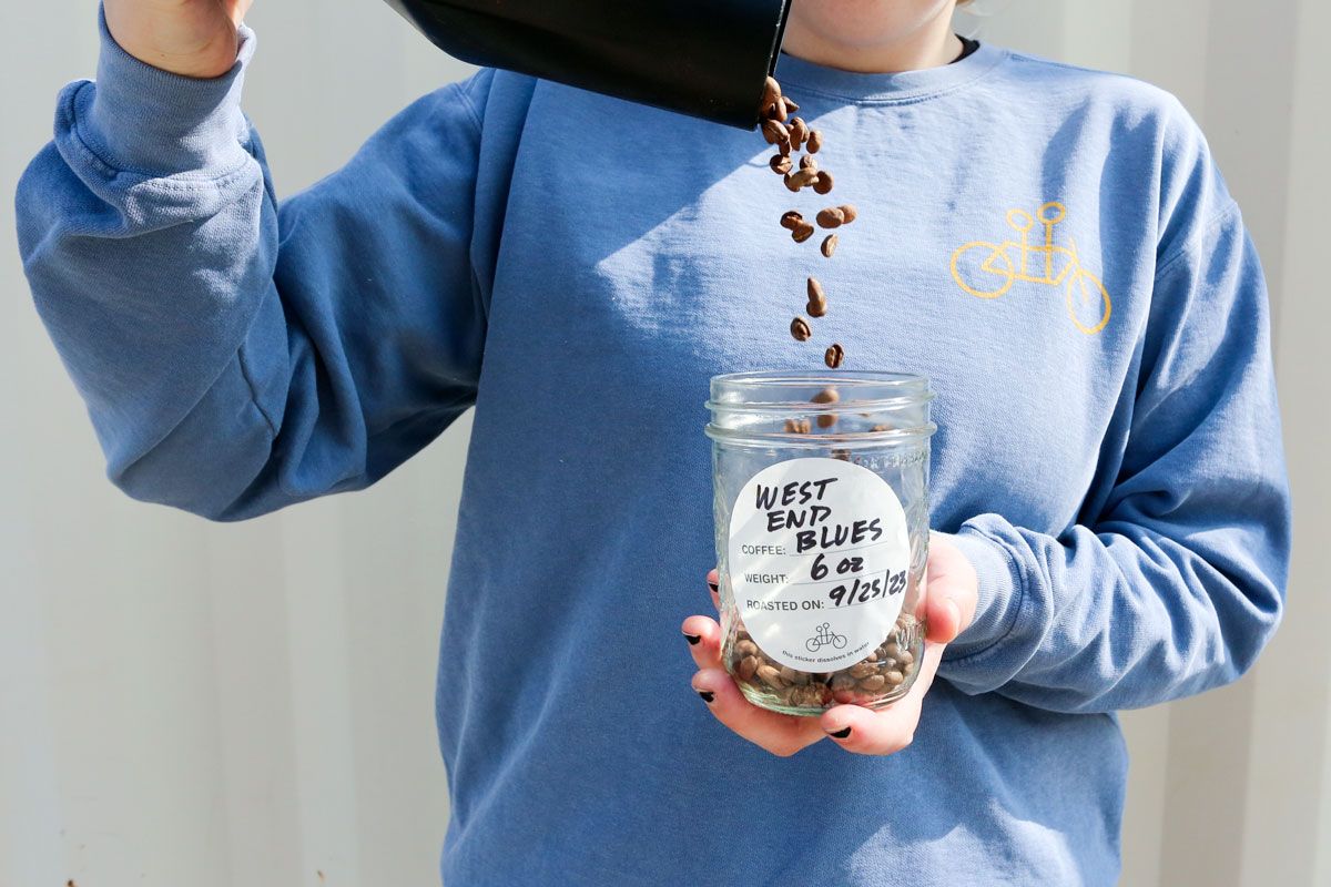 Person in a blue sweatshirt pouring coffee beans into a glass jar labeled "west end coffee blues" held in their hands against a white wall background.