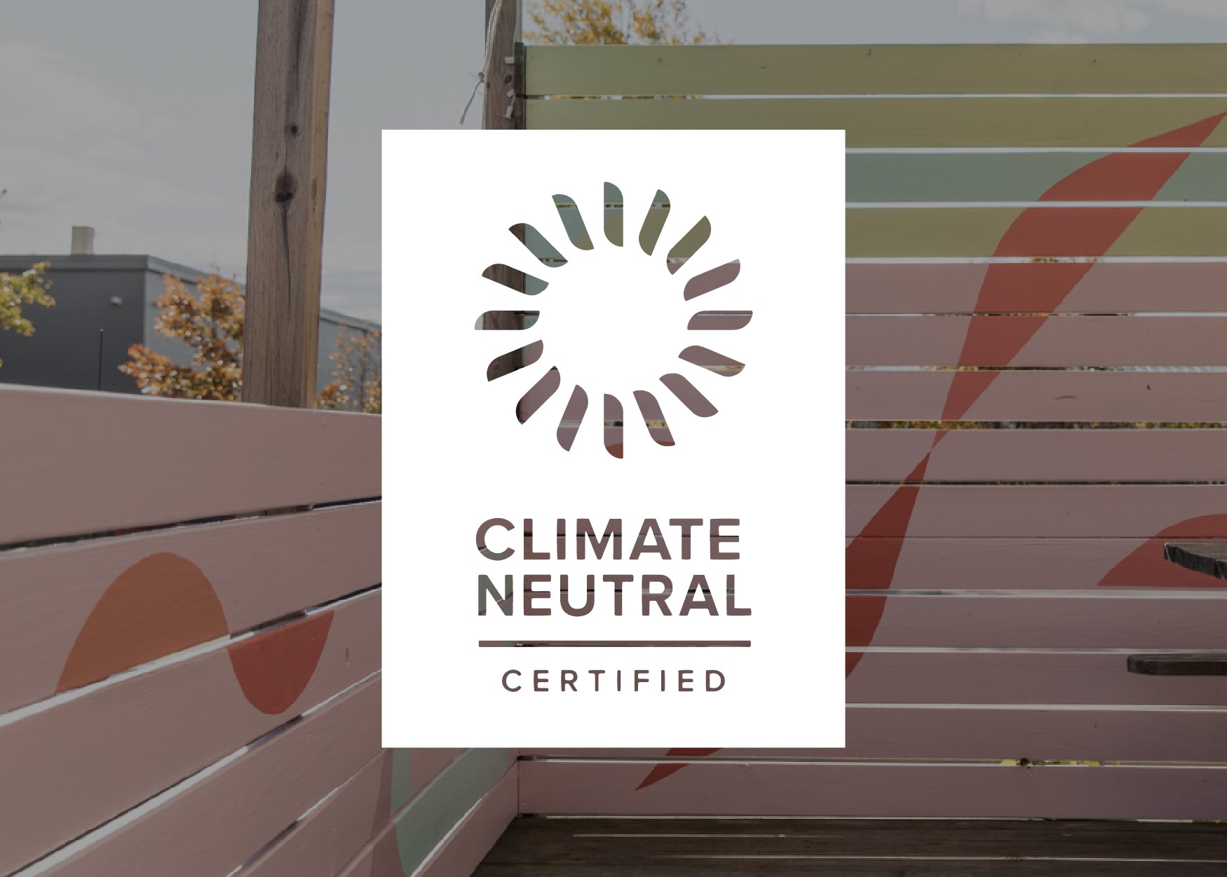 A poster reading "climate neutral certified" with a circular logo, mounted on a wooden bench painted with pastel stripes, against a backdrop of trees and a cloudy sky.