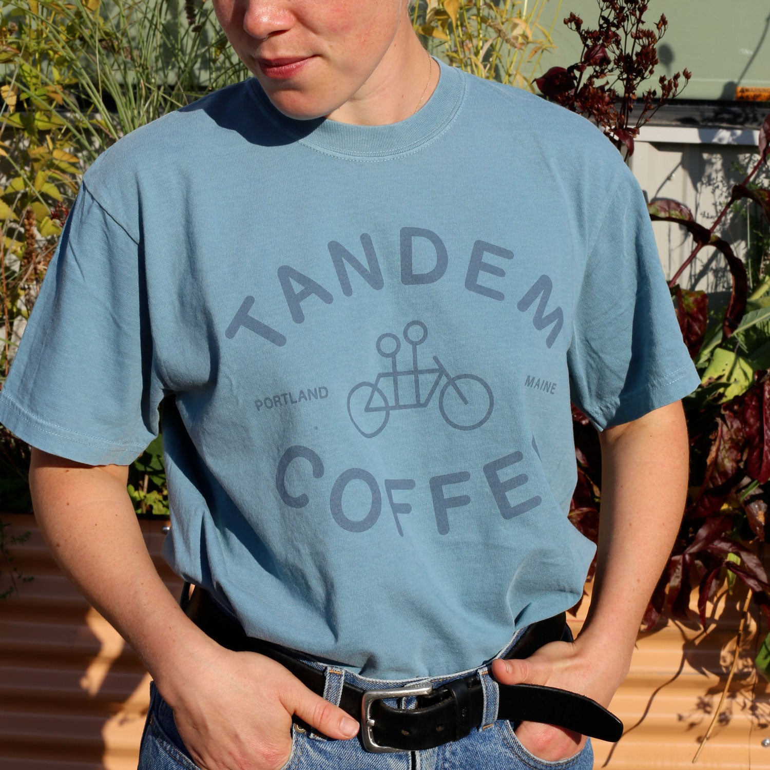 A person wears the Ice Blue Logo Tee from Tandem Coffee Roasters, featuring a bicycle graphic, standing outdoors with hands in pockets amid greenery.