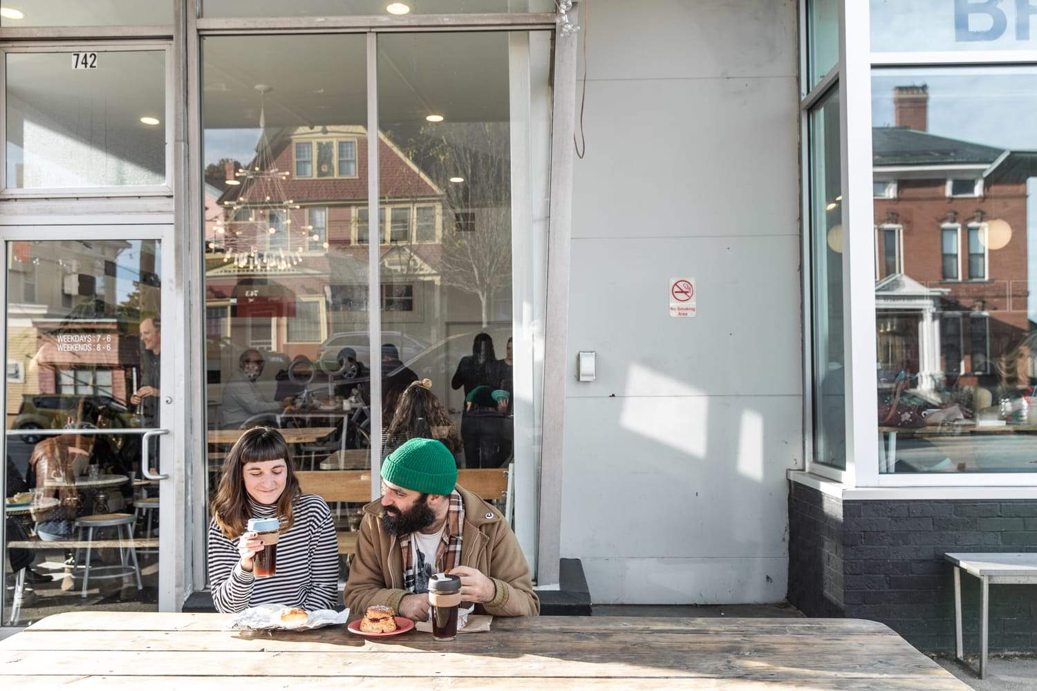 A man and woman sit at a wooden table outside a café in Portland, Maine. They are enjoying coffee and pastries from Tandem Coffee while smiling and chatting. Behind them, large windows reveal people inside the café. Sunlight illuminates the scene, and houses are visible across the street.