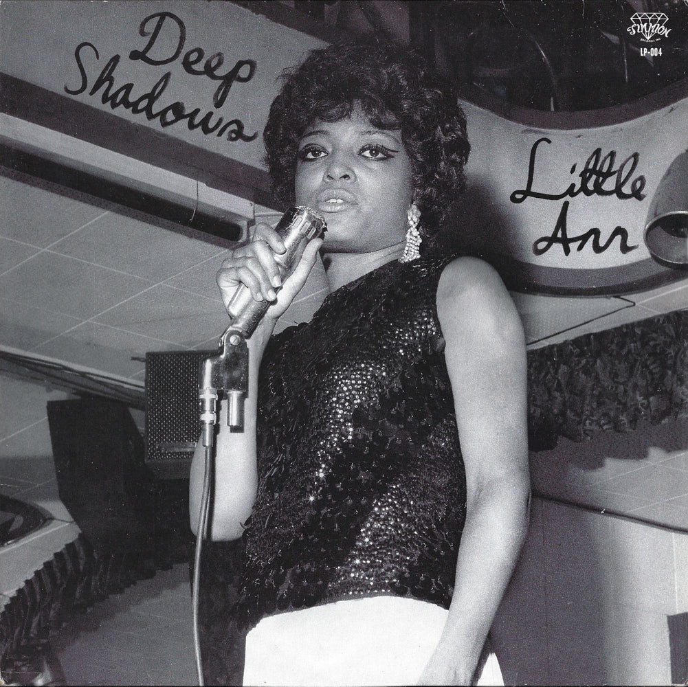 Black and white photo of a woman singing into a microphone. She has an afro hairstyle and wears a sequined top and earrings. Signs read "Tandem Coffee Roasters - Little Ann's Deep Shadows" and "streaming".