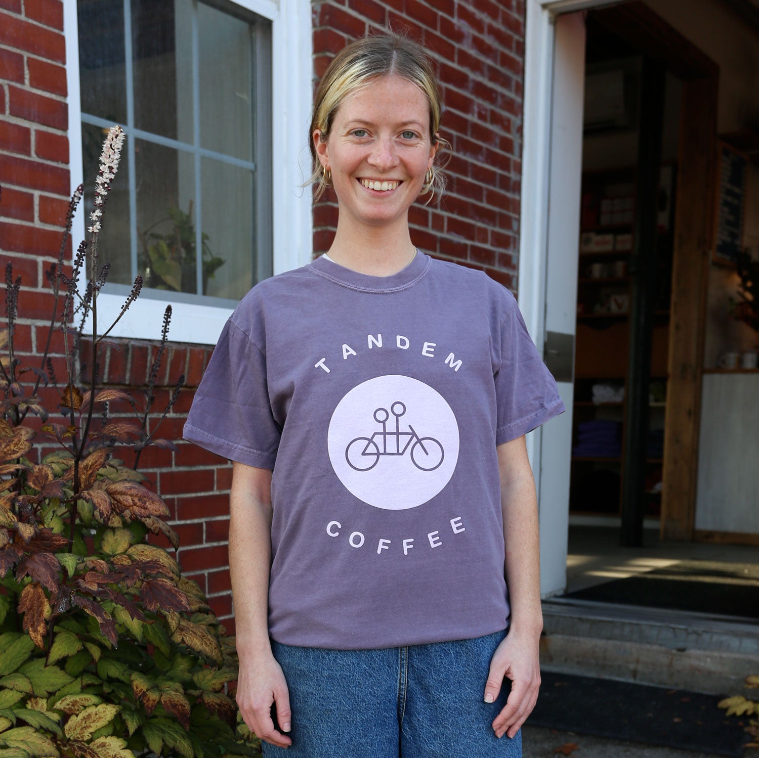A person stands smiling in front of a brick building, wearing a "Purple Circle Logo Tee" by Tandem Coffee Roasters, adorned with a tandem bicycle graphic. The door is open, offering a peek inside. Vibrant plants add color to the scene on the left.