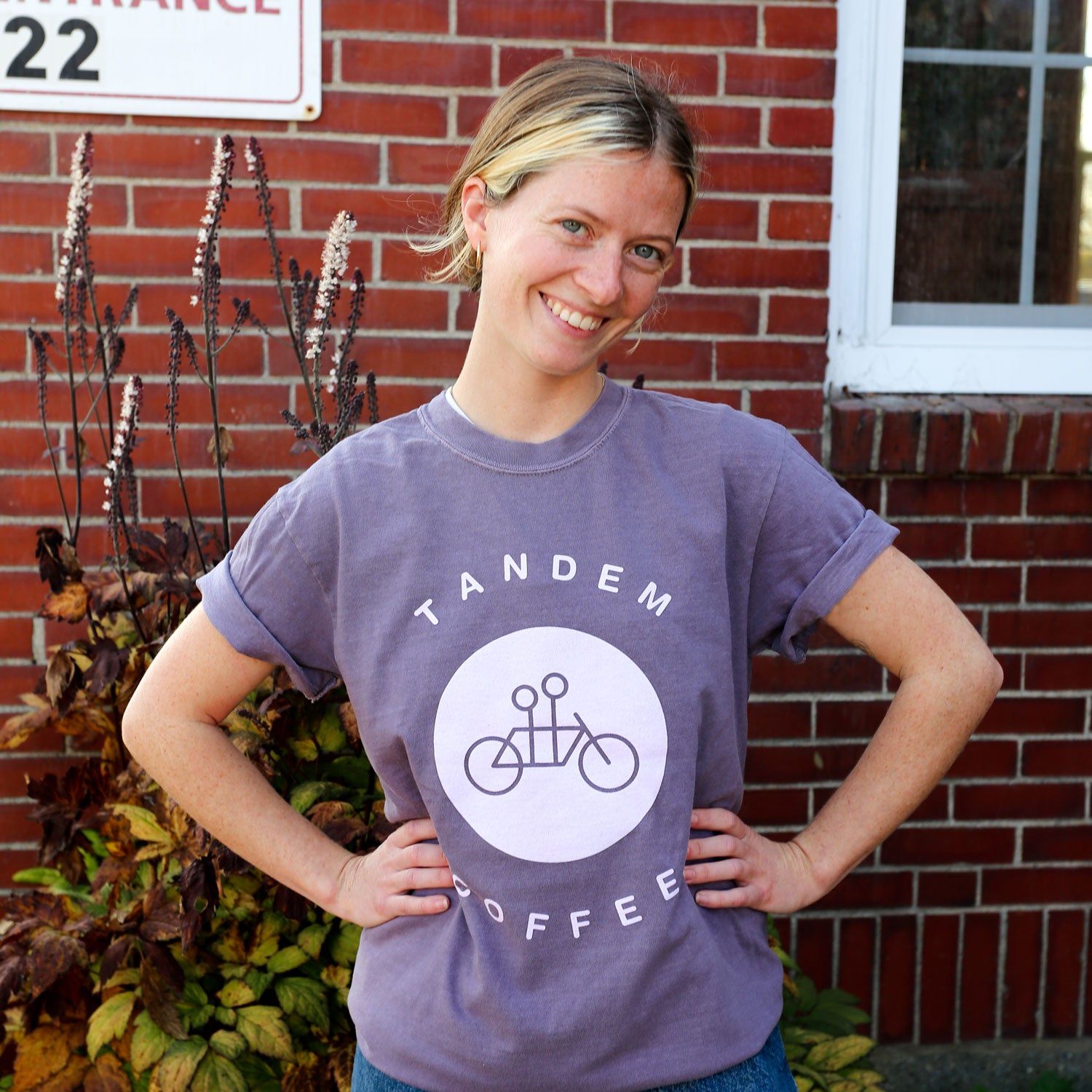 A person wearing a "Purple Circle Logo Tee" from Tandem Coffee Roasters, celebrated for its Comfort Colors softness, poses with hands on hips. They stand smiling at the camera in front of a brick wall adorned with nearby plants.