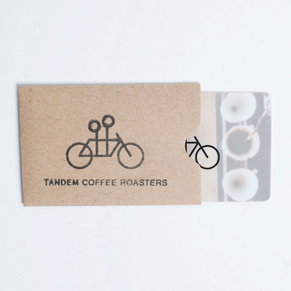 Two physical gift cards for Tandem Bakery, one with a tandem bicycle and logo etched on a cardboard texture, the other featuring a coffee bean design on a translucent background.
