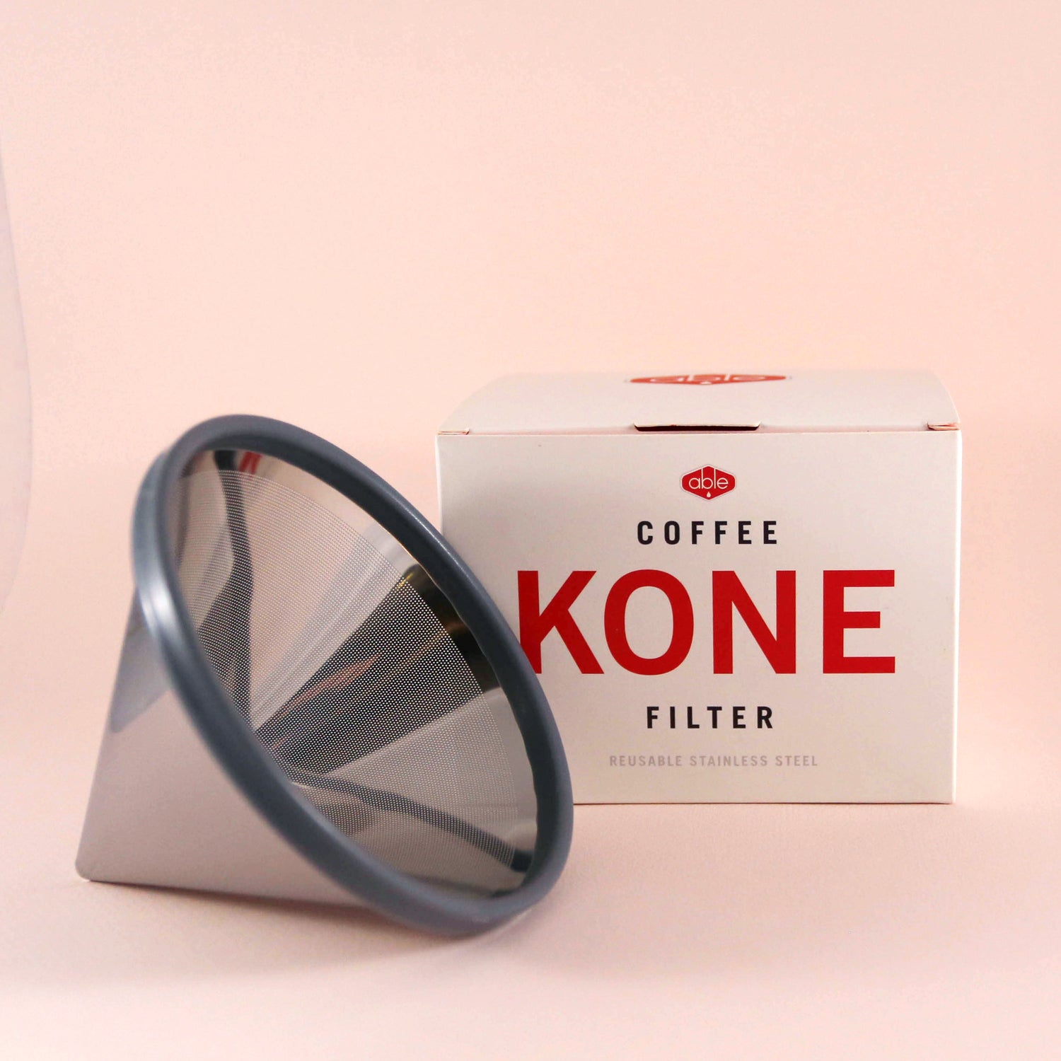A Tandem Coffee Roasters Able Coffee Kone stainless steel reusable coffee filter next to its packaging box on a light pink background. The box displays the product name prominently.