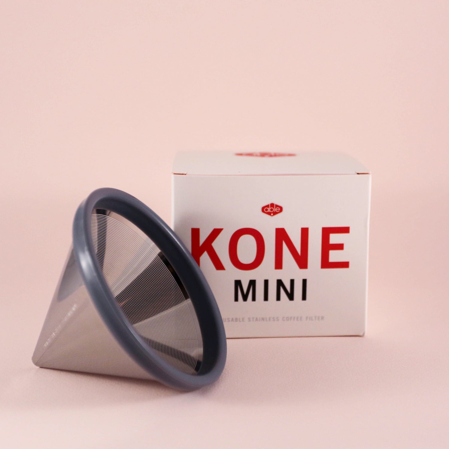 A gray, cone-shaped reusable stainless steel filter titled "Tandem Coffee Kone Mini" leaning against its packaging box on a pink background.