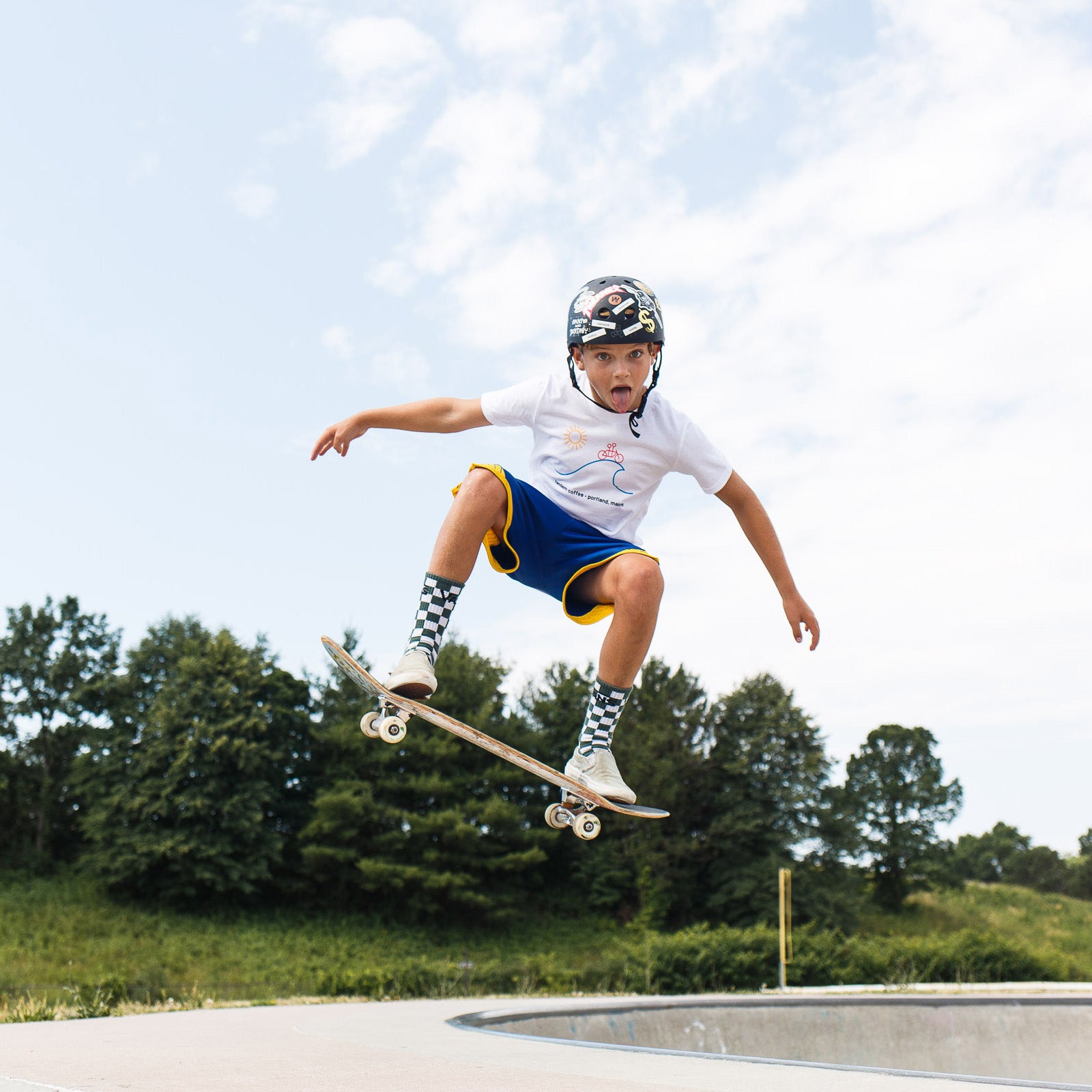 A young boy wearing a helmet, knee pads, and a Tandem Kids Tandem Sun Tee performs a mid-air skateboarding trick at a skatepark under a clear sky.