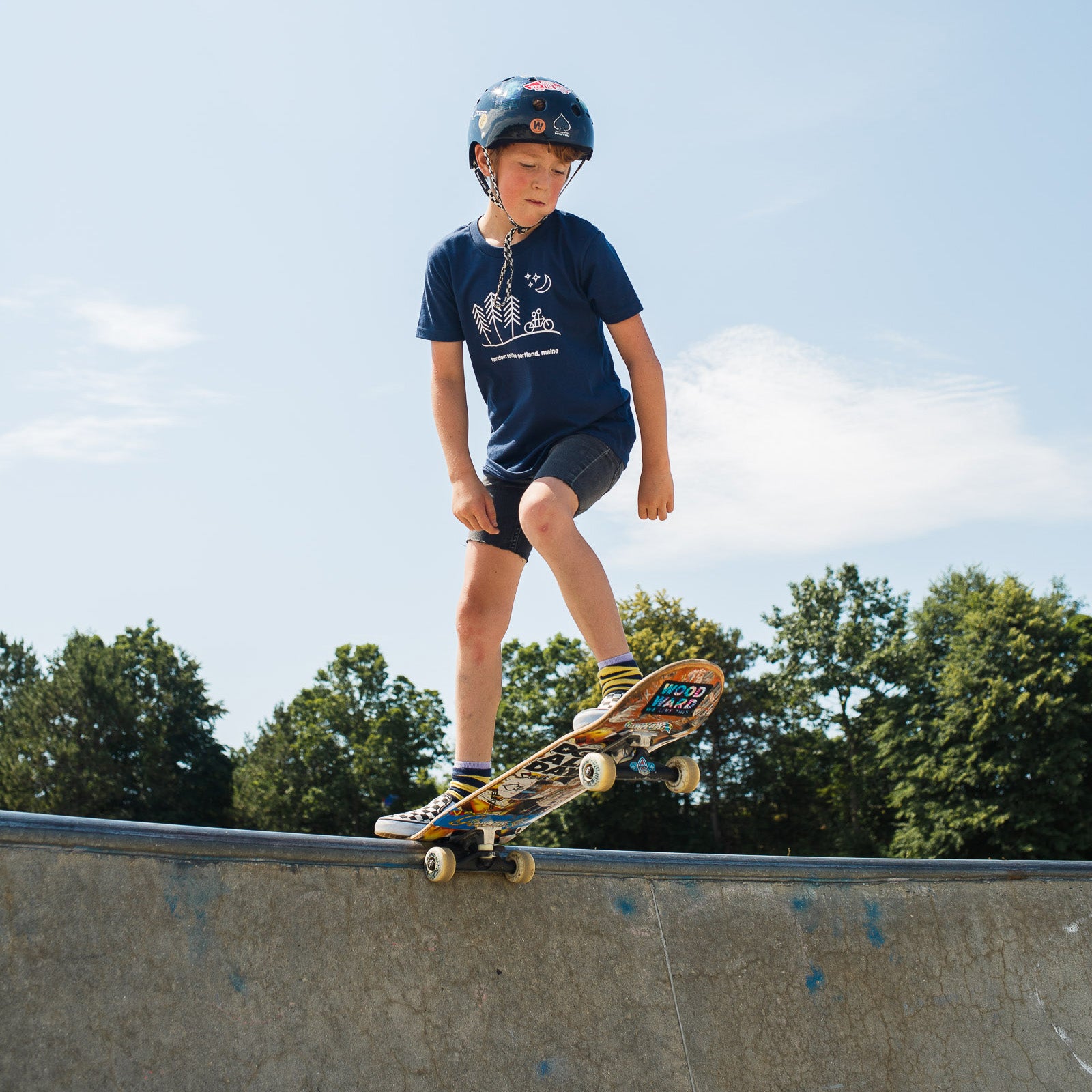 A young skateboarder in a helmet and 100% cotton Tandem Kids Tee stands confidently with a skateboard, looking off into the distance, with another person in the background at a skate park.