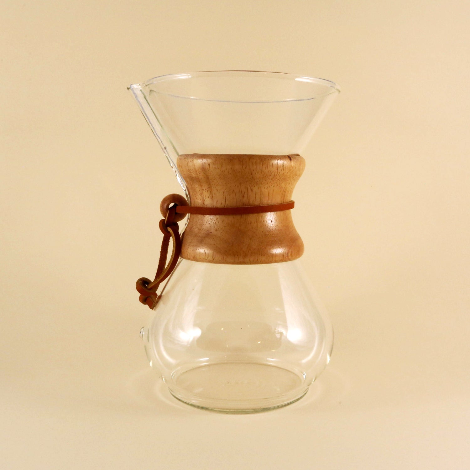 Tandem Coffee Roasters' Chemex 6 Cup Brewer, a clear glass coffee maker with a reusable filter, conical top, and bulbous bottom, featuring a wooden collar and leather tie against a beige background.