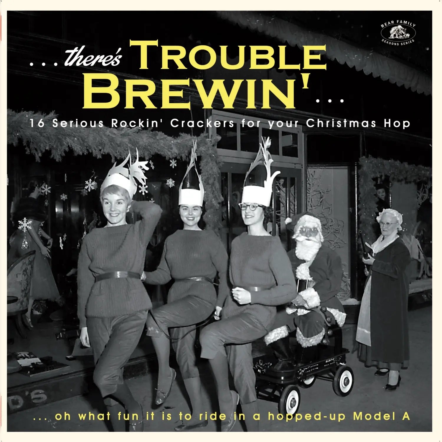 Three women dressed in festive outfits and Santa hats pose with a mini car and a Santa figure in a holiday-themed setting. The text on the image says, "...there's Trouble Brewin'..." highlighting "16 Serious Crackers for your Christmas Hop" featuring hits like "Rock Around The Christmas Tree." This product is brought to you by Tandem Coffee Roasters.