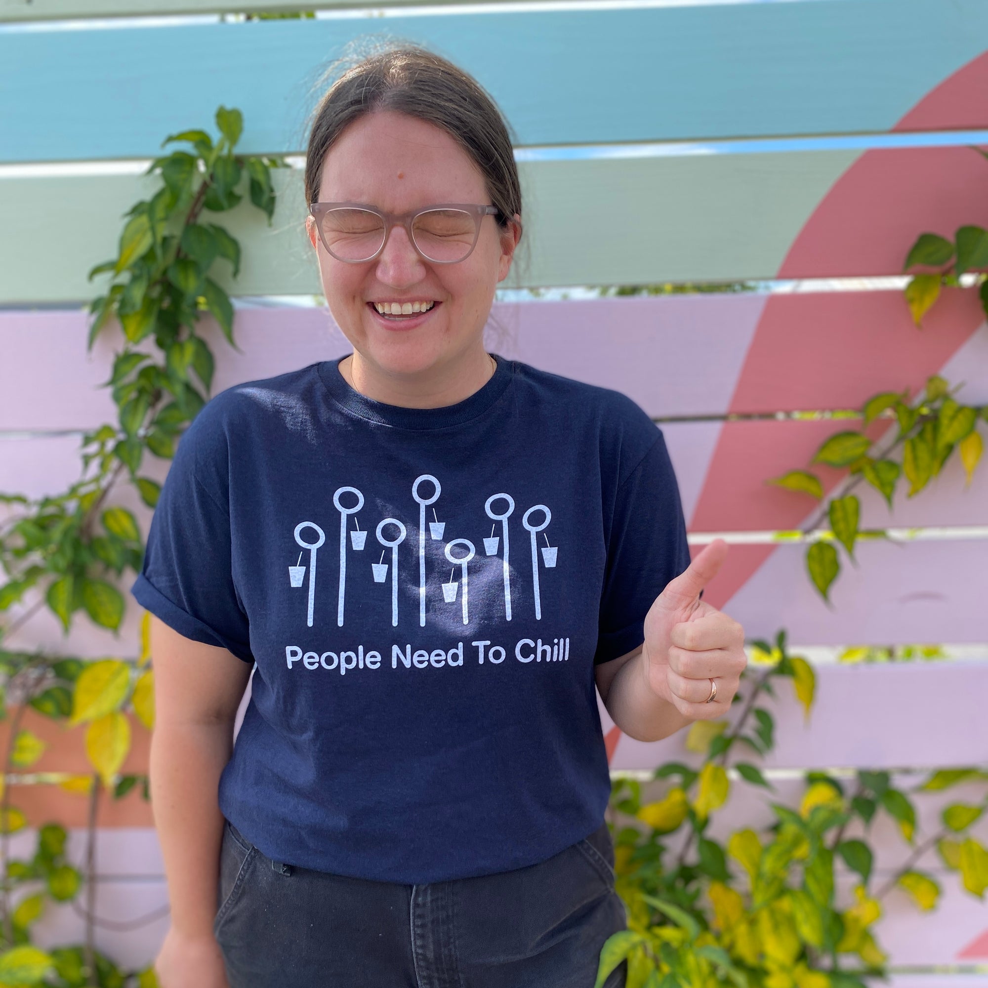 A young woman in glasses, smiling and giving a thumbs up, wearing a navy blue Tandem Coffee Roasters "People Need To Chill" Tee in front of a colorful striped background with greenery.