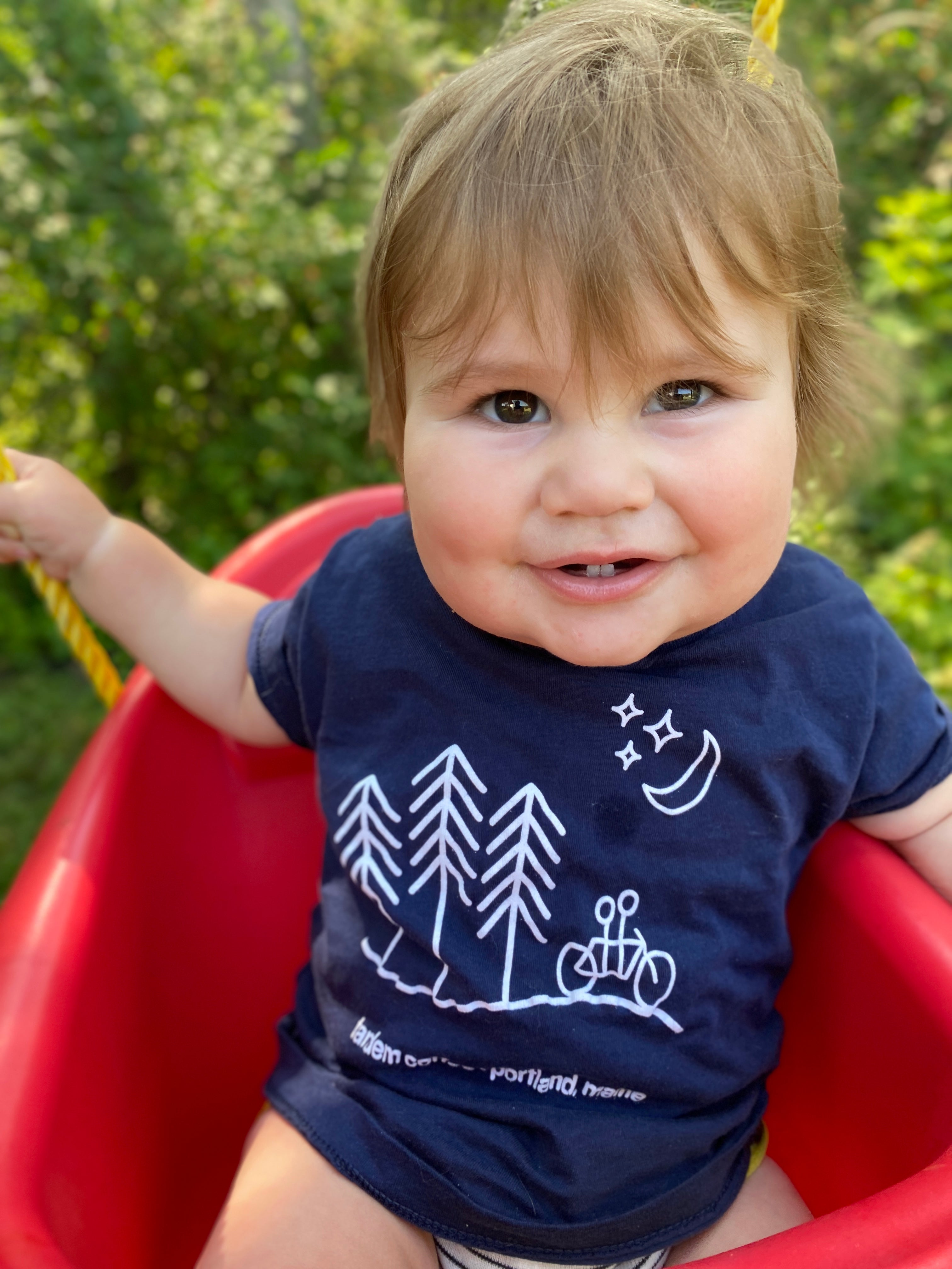 A joyful toddler with light brown hair sitting in a red swing, wearing a dark blue Tandem Kids Tandem Moon Tee with tree and moon graphics. The background is lush and green.