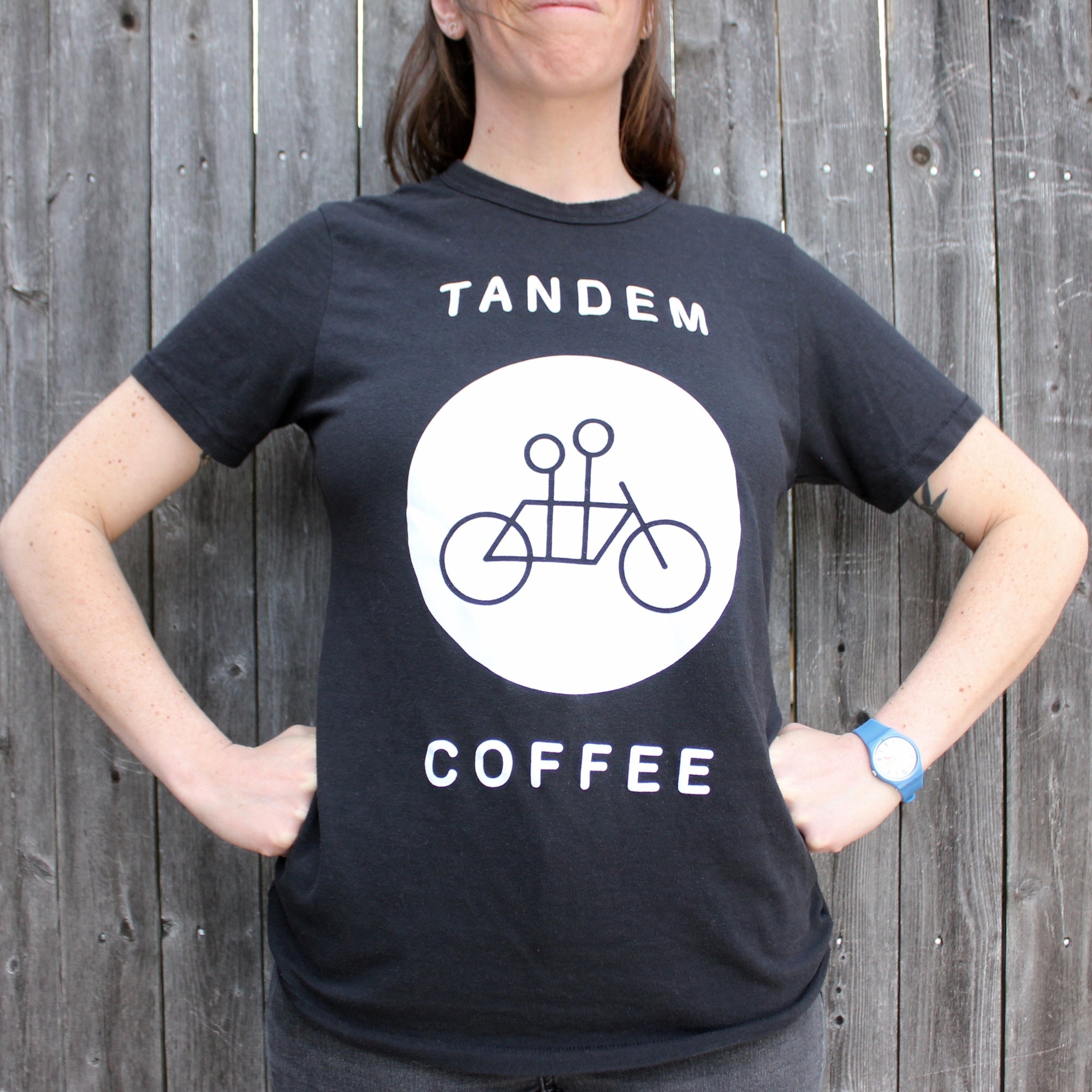 A woman in a black Tandem Coffee Roasters Circle Logo Tee featuring the words "tandem coffee" and a graphic of a tandem bicycle stands against a wooden backdrop.