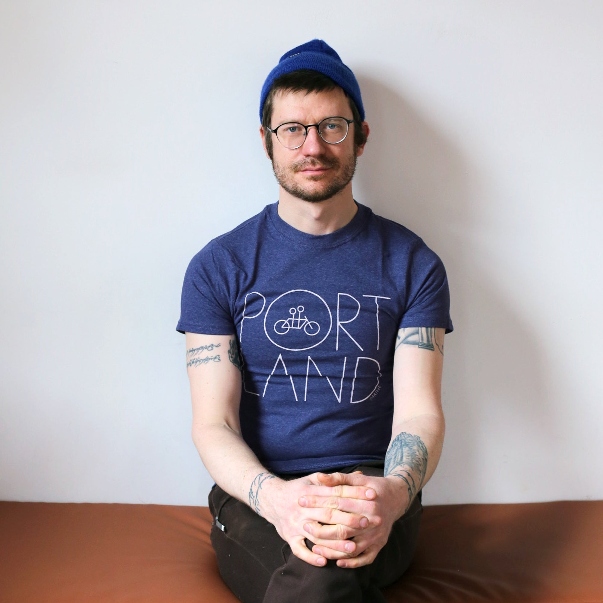 A man wearing a blue beanie and a Recycled Portland Tee from Tandem Coffee Roasters sits cross-legged on a brown surface against a white wall, looking at the camera.
