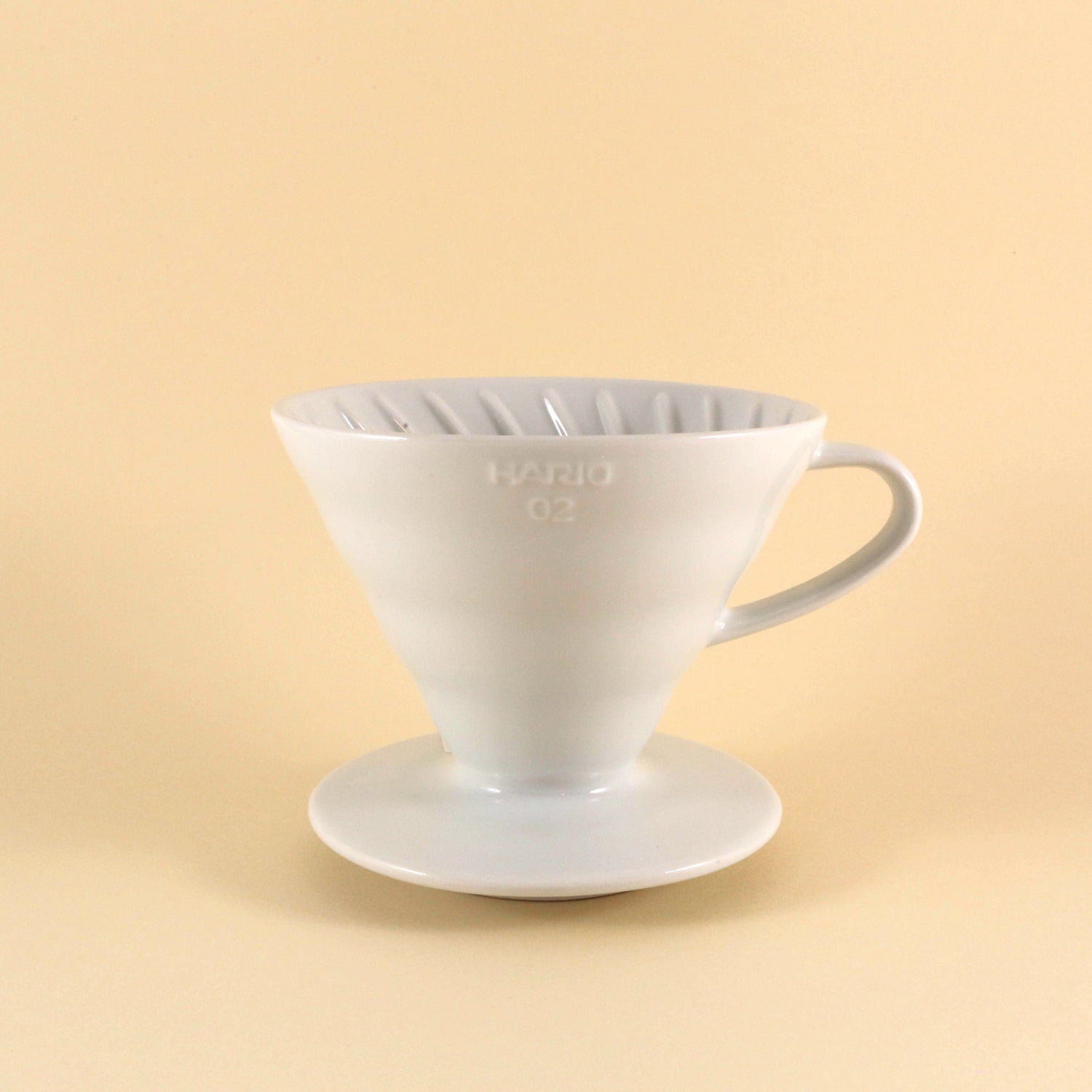 A white vendor-unknown Hario Ceramic V60 Dripper with a reusable filter sitting on a matching saucer against a soft beige background.