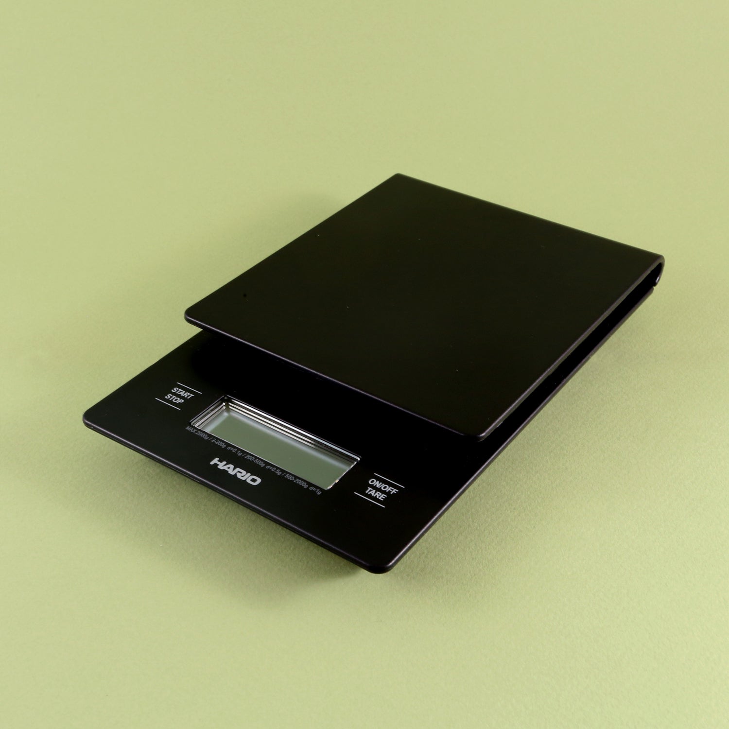 A black Hario V60 Drip Scale with a visible LCD display, sitting on a pale green background. The scale is compact and has a sleek, modern design, ideal as a home coffee scale.