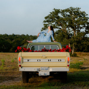 A woman in a long blue dress sits atop a vintage yellow Ford pickup filled with red flowers, parked in a grassy field near trees, with the Waxahatchee - Saint Cloud coffee by Tandem Coffee Roasters featured.