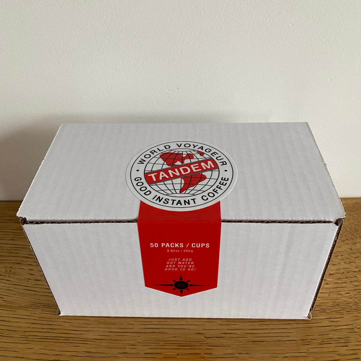 A white cardboard box with red and white labels, featuring the text "Tandem Coffee Roasters World Voyageur instant coffee," plus product details like "50 packets/cups." The box sits on a wooden surface.