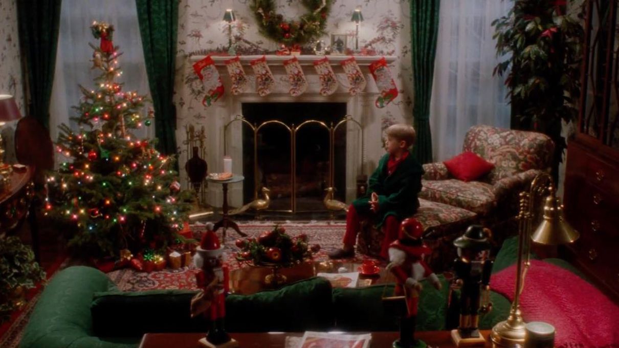 A cozy living room decorated for Christmas, with a lit tree, stockings on the fireplace, garlands, and a young boy in a green robe sitting on a couch, gazing at the festive decorations.