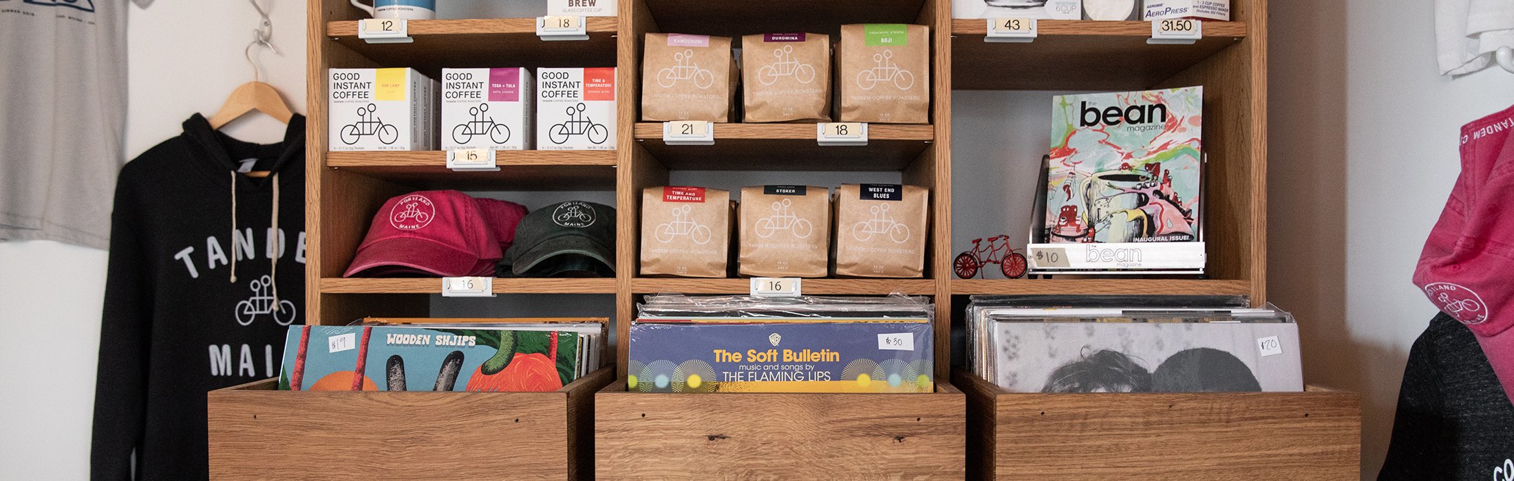 Wooden shelves displaying coffee bags, assorted hats, magazines, and records. Tandem-branded merchandise includes t-shirts, a sweatshirt, and a pink bandana. Price tags are visible on most items.