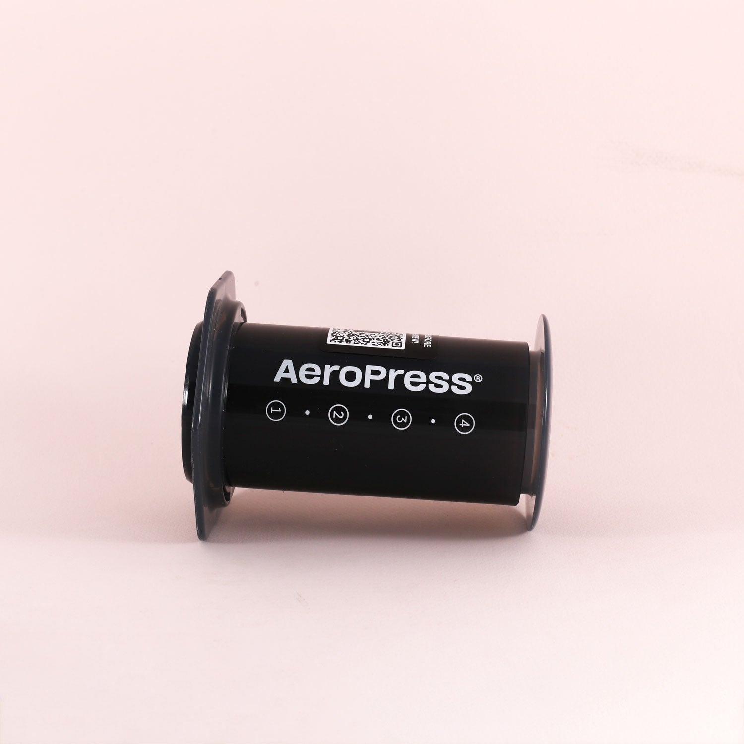 A black Aeropress coffee maker by Tandem rests on its side against a light pink background, displaying the brand name and simple white icons along the side.