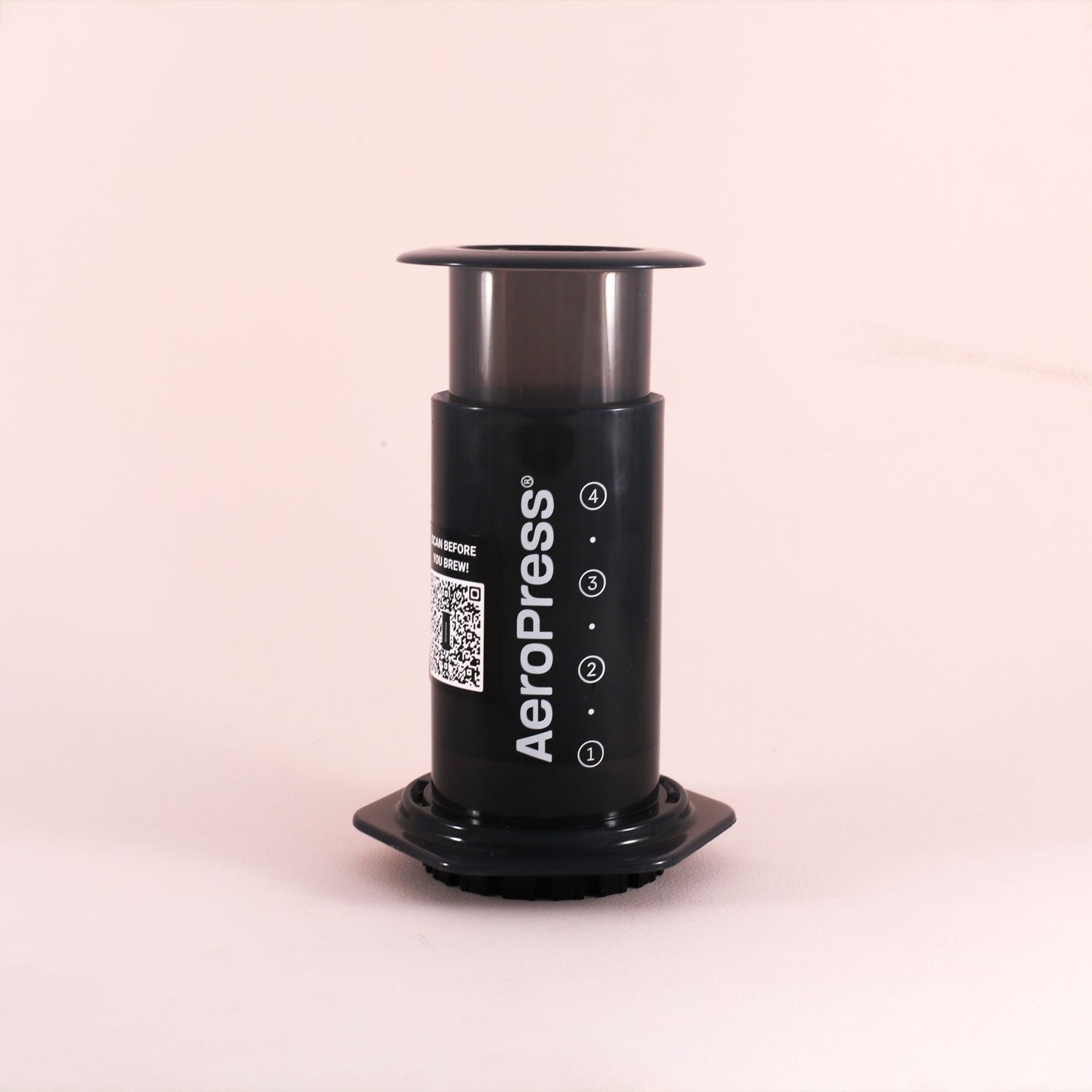 A black Aeropress coffee maker by Tandem stands upright on a light pink surface, with the Tandem brand name displayed vertically along the side.