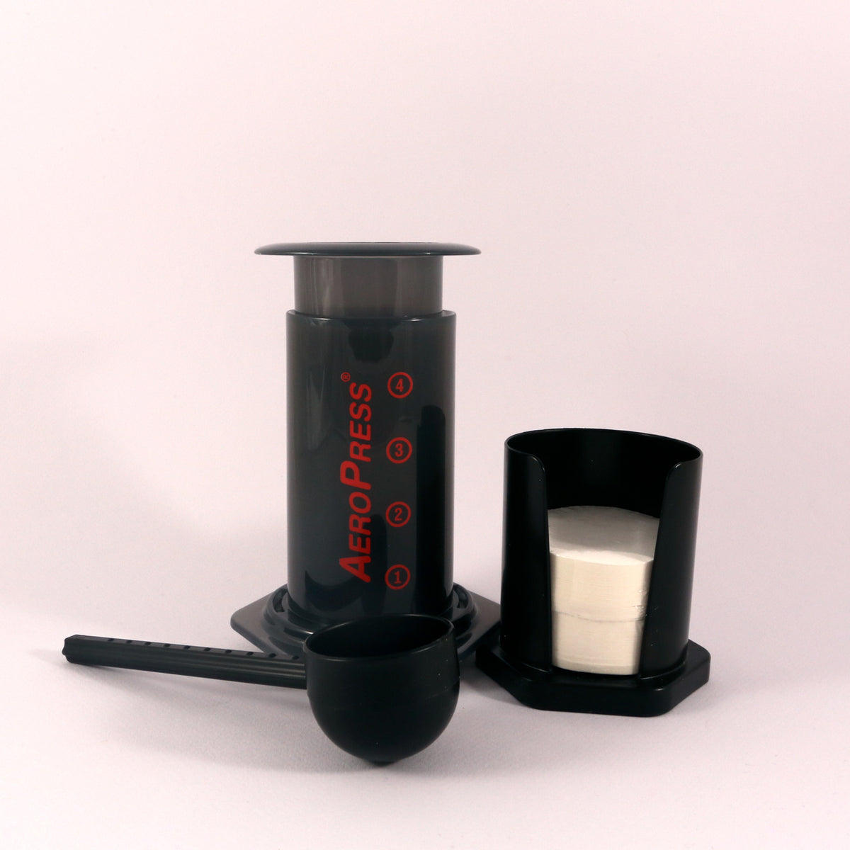 An Tandem aeropress coffee maker, touted as the world's greatest, with its components arranged neatly: the press, filter cap, scoop, and black funnel, against a light gray background.