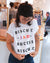 A woman in a cafe adjusts her Loaded Biscuit Tee from Tandem Coffee Roasters, with glasses and tattoos visible, and other patrons in the background.