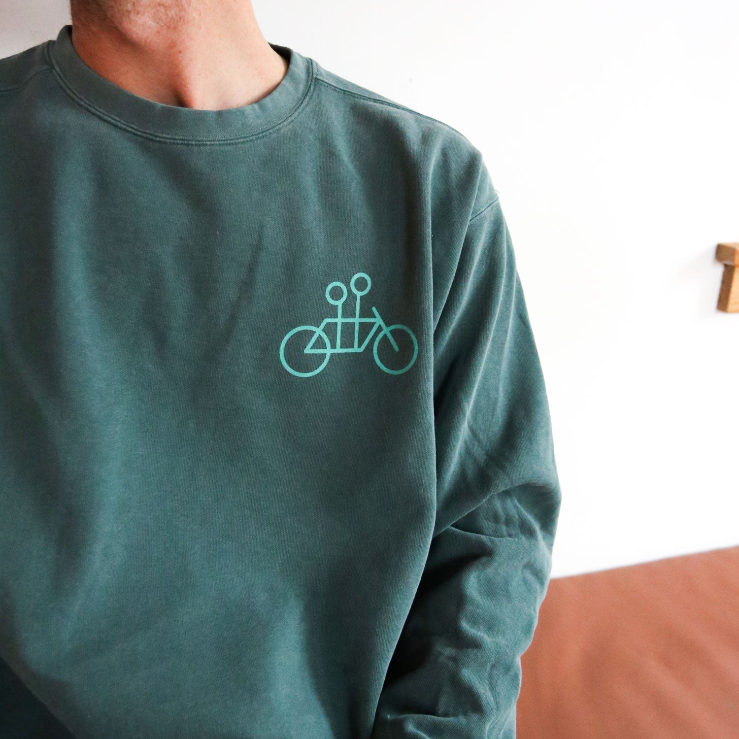 A person wearing a Blue Spruce Tandem Crewneck by Tandem Coffee Roasters, featuring a minimalist bicycle design, stands in front of a white and brown background, embodying the cozy vibe often associated with Comfort Colors attire.