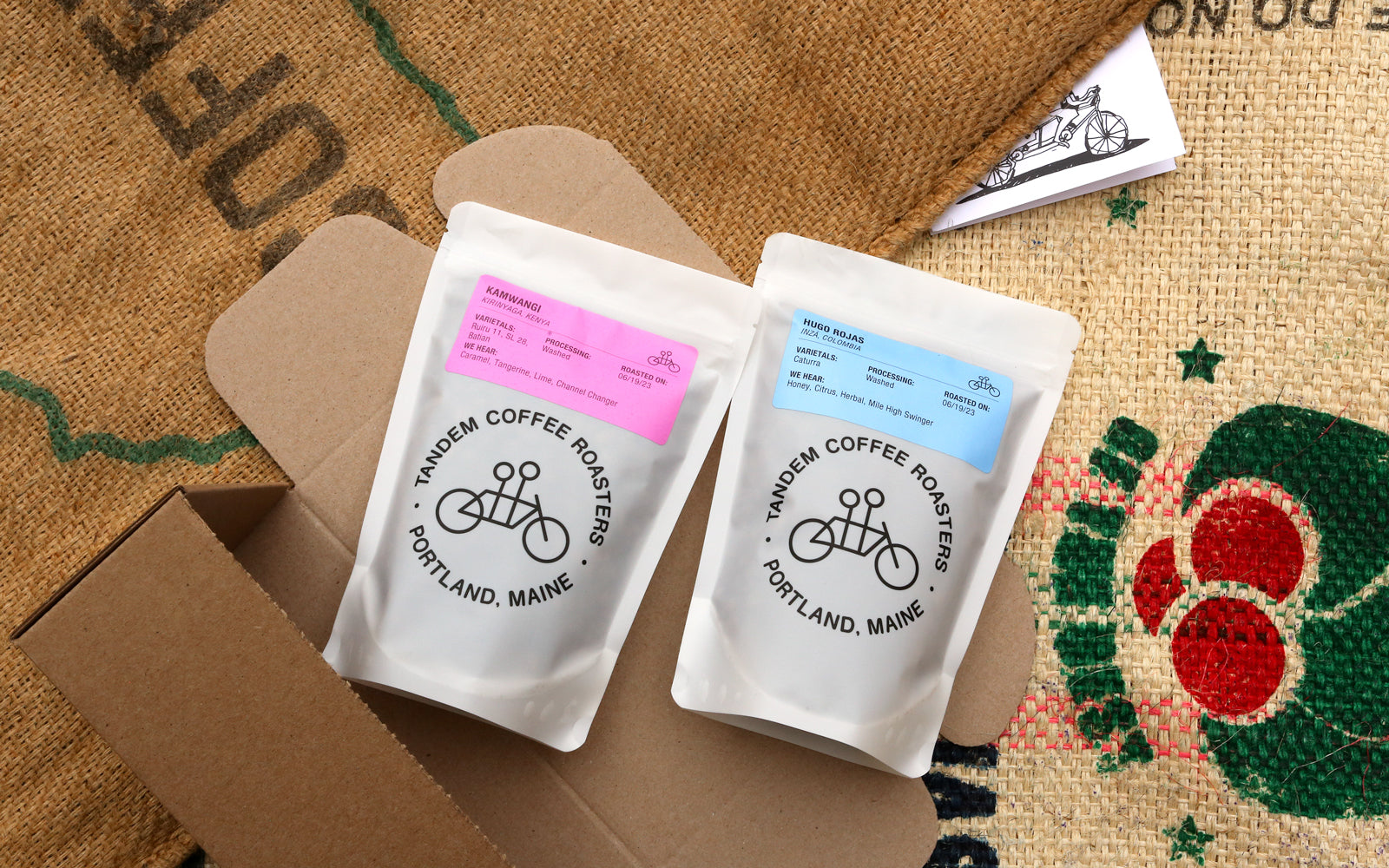 Two coffee bags labeled "warden coffee roasters - portland, maine" with pink and blue labels on a burlap surface with a cardboard box and floral pattern.