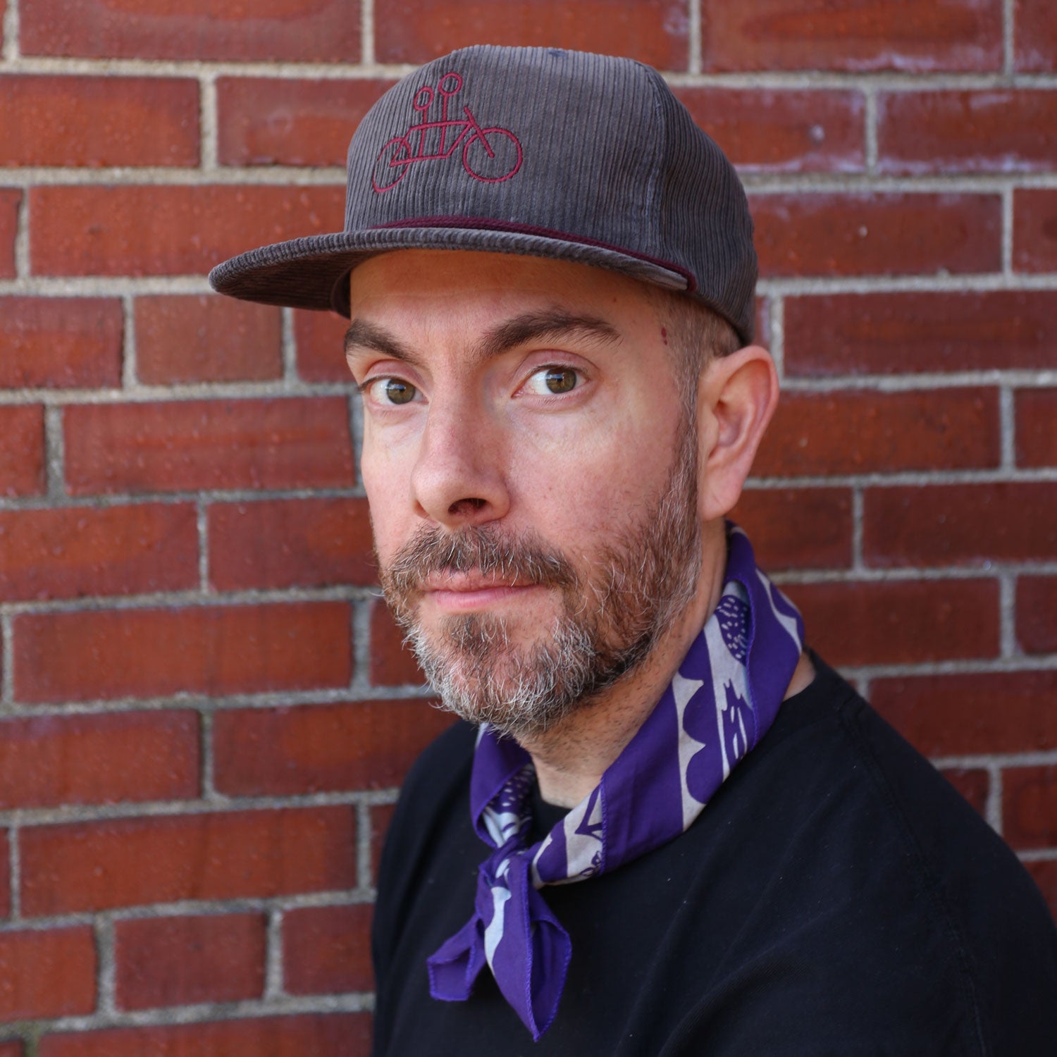 A bearded person wearing a gray Corduroy Tandem Hat from Tandem Coffee Roasters, featuring a bicycle design, and a purple bandana around their neck stands in front of a red brick wall, looking into the camera.