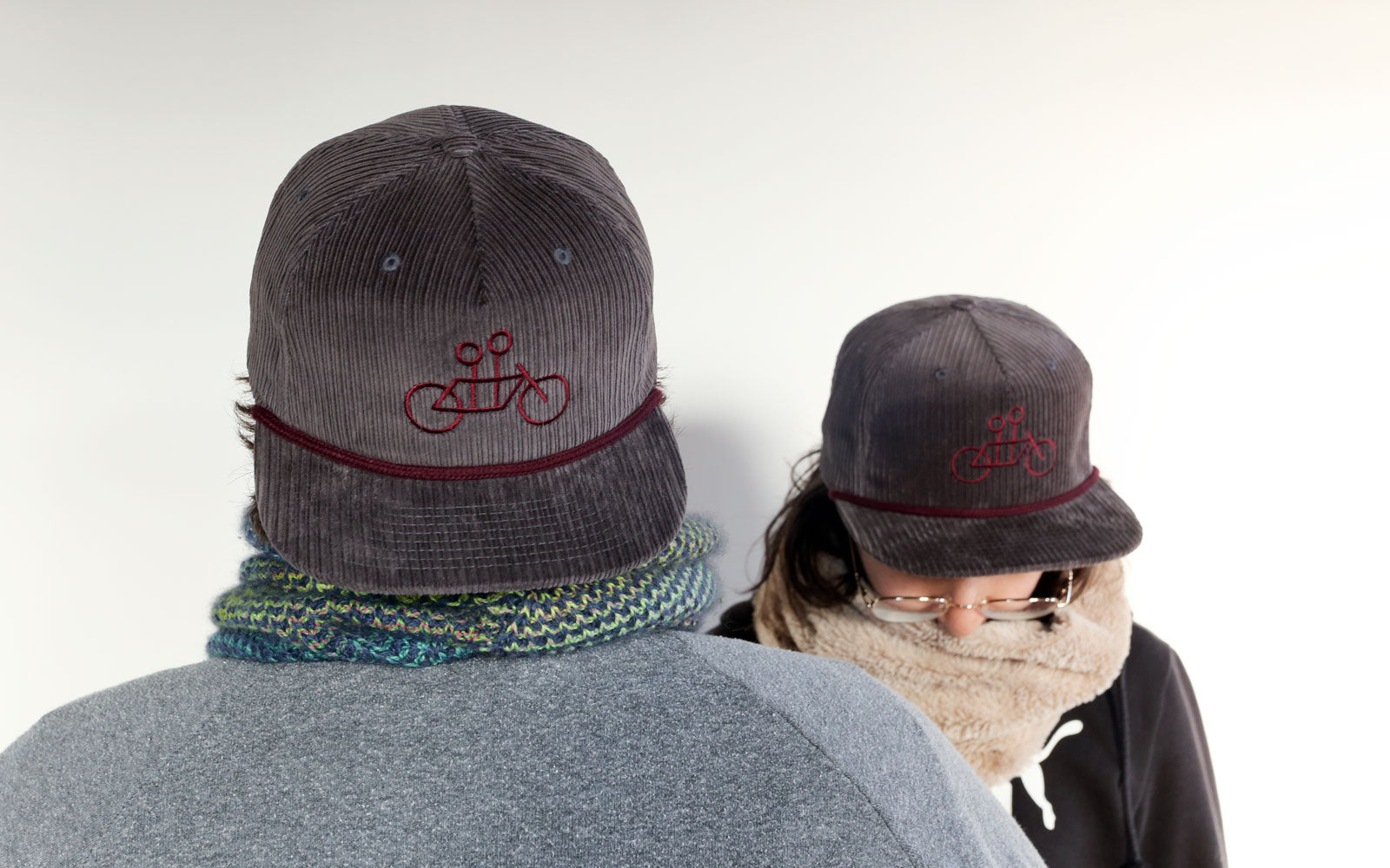 Two people from behind, wearing dark hats embroidered with a red bicycle logo. one person, likely female, wears glasses and a scarf, while the other, likely male, sports a knitted multicolored scarf.